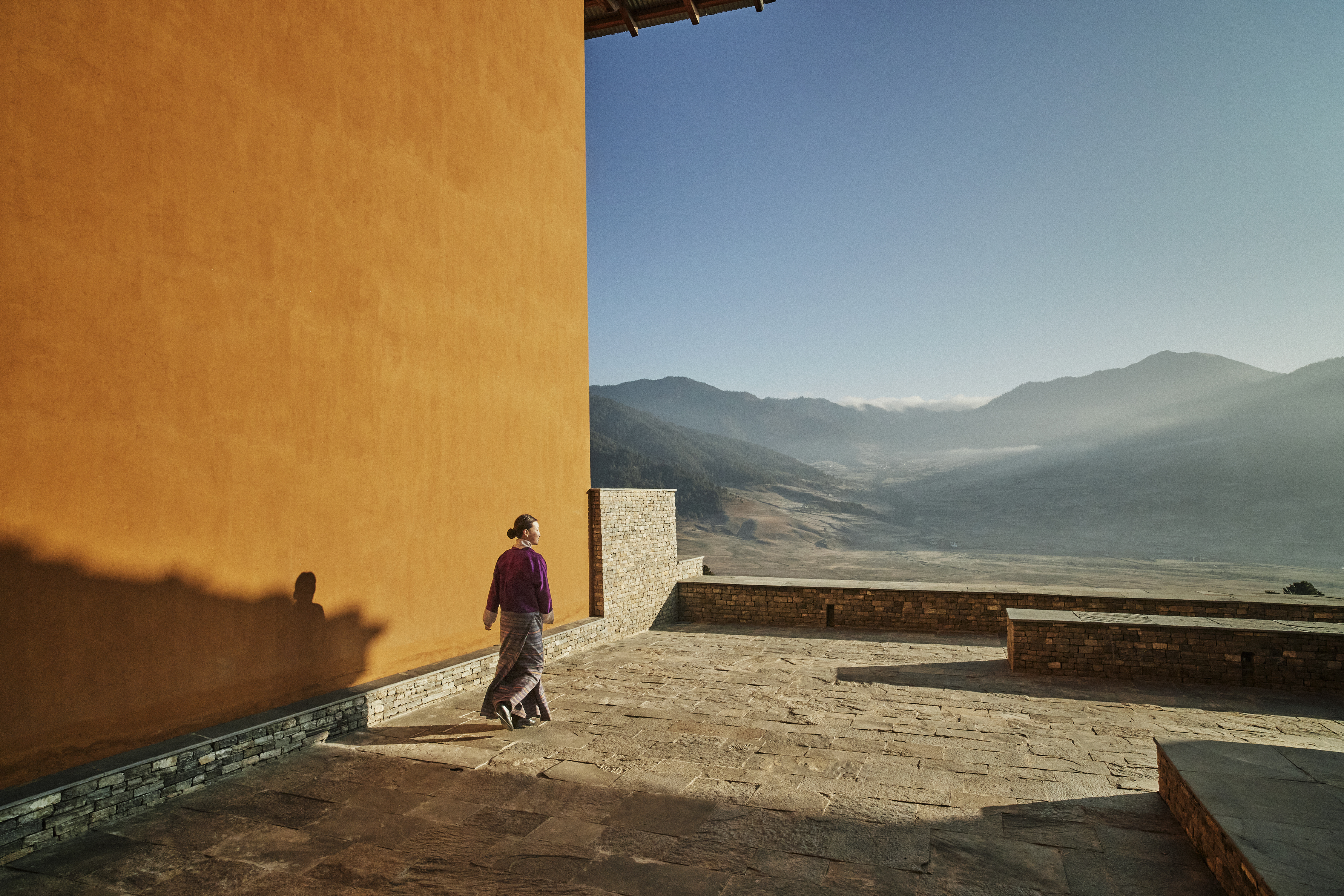 A traditional Bhutanese lodge courtyard with stone walls, wooden balconies, and a welcoming figure.