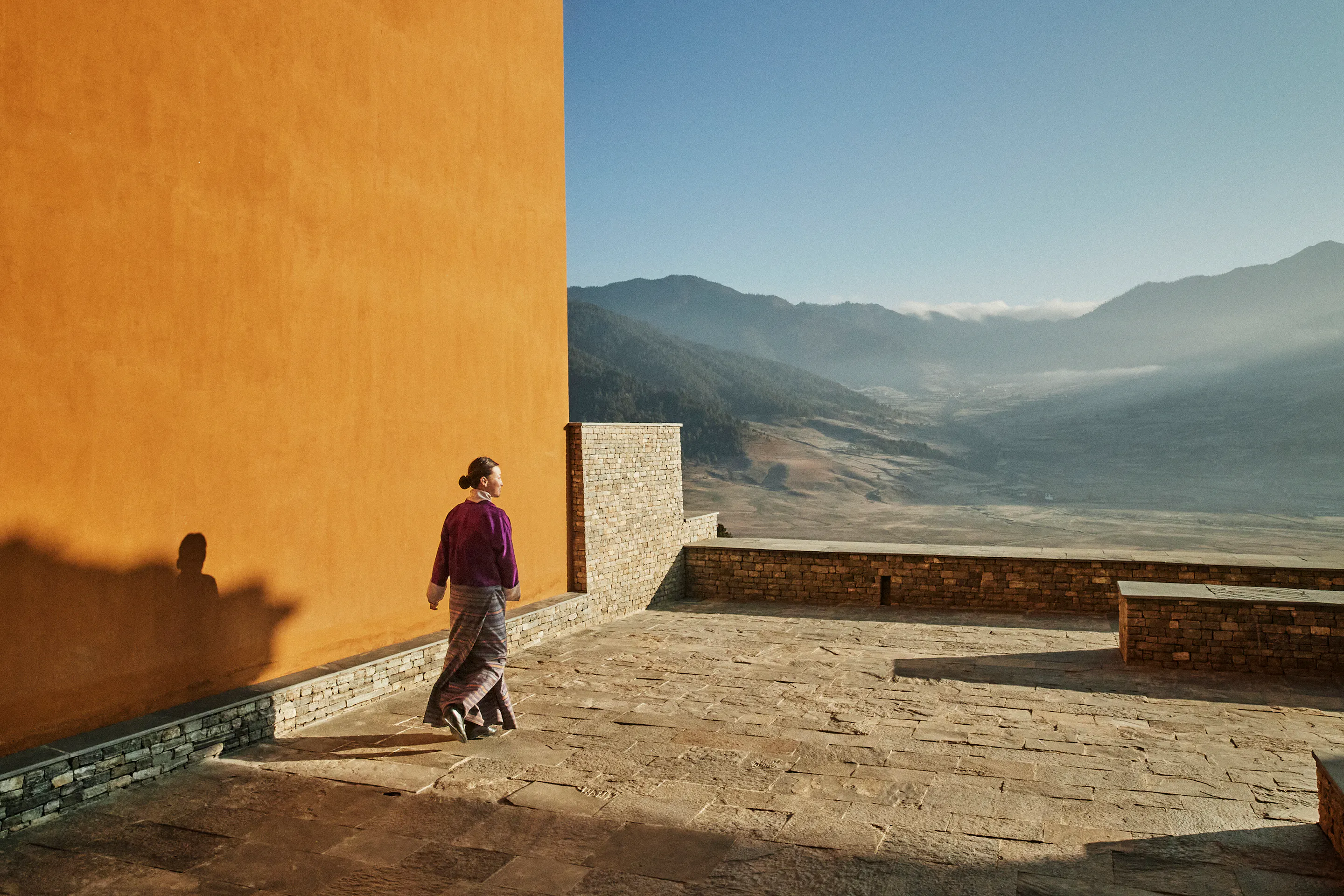 A traditional Bhutanese lodge courtyard with stone walls, wooden balconies, and a welcoming figure.