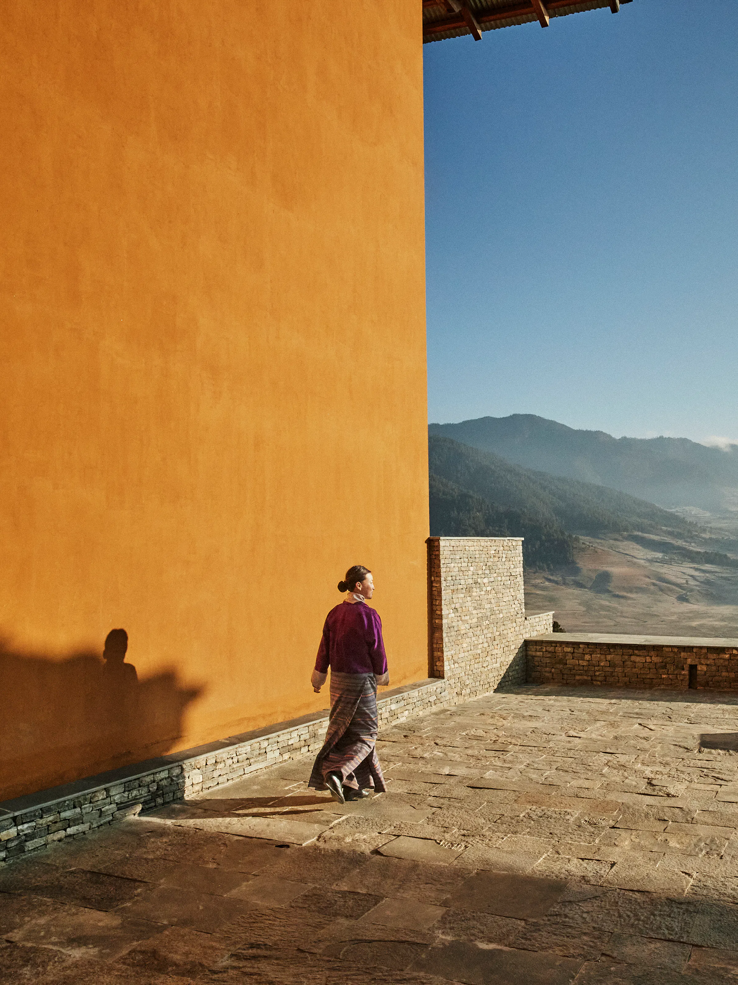 A traditional Bhutanese lodge courtyard with stone walls, wooden balconies, and a welcoming figure.