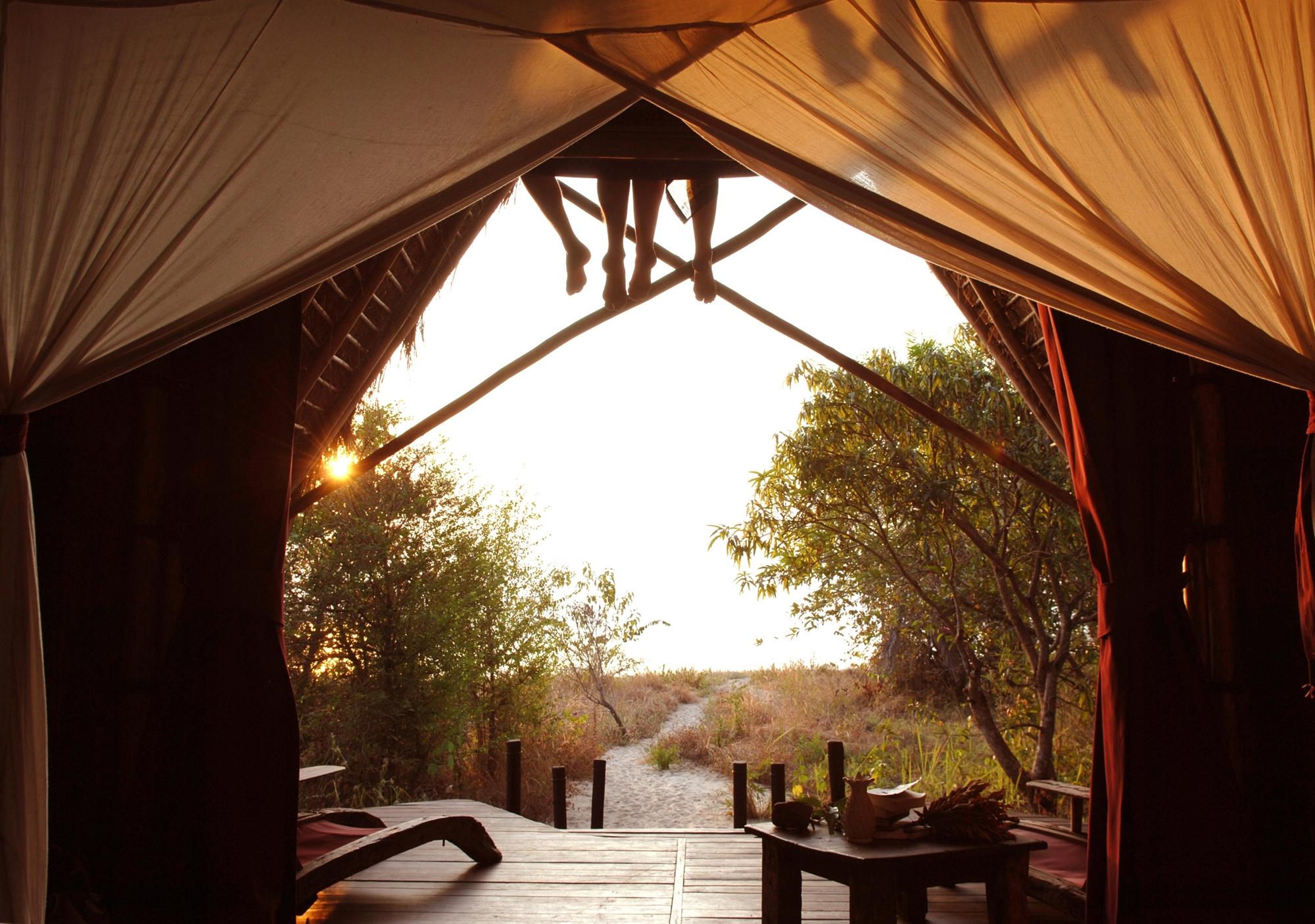 An open-sided canvas tent lodge overlooks Lake Tanganyika through palm fronds at golden hour.