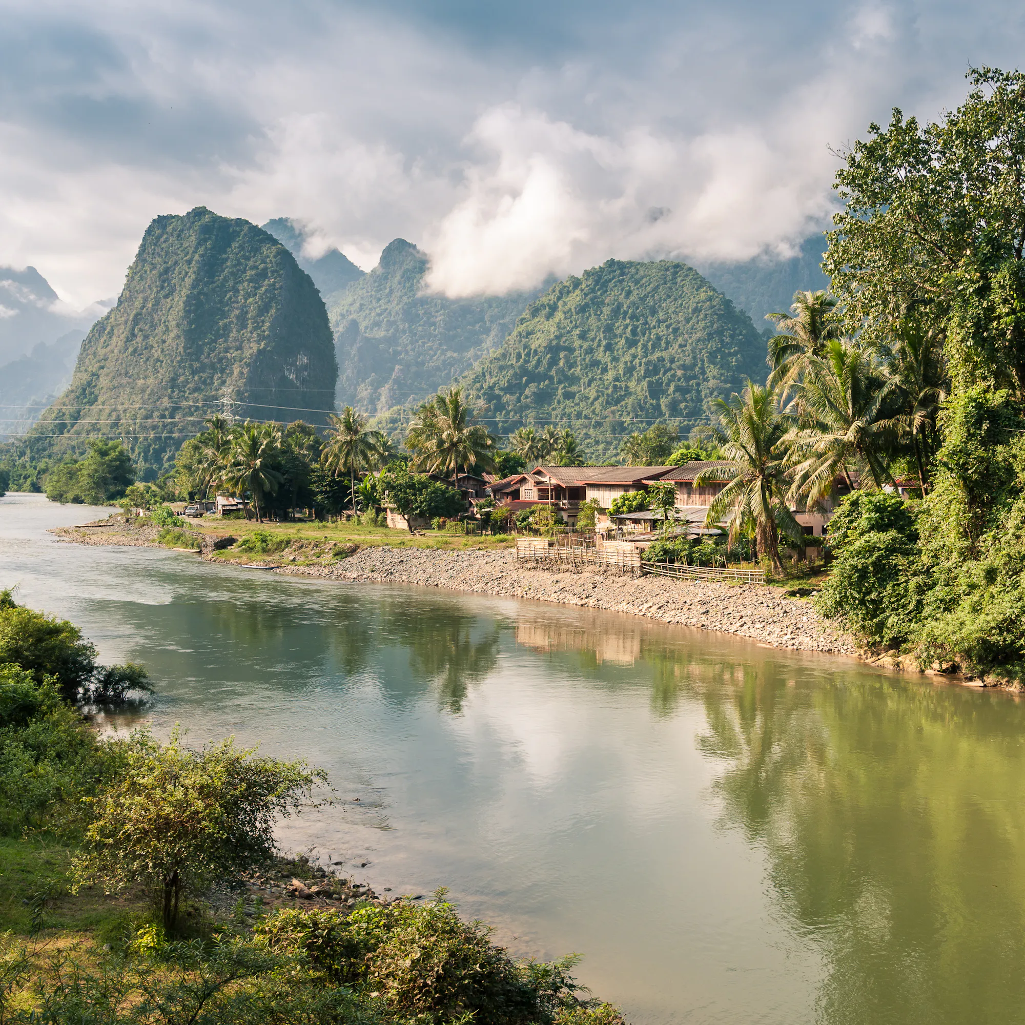 A flat green river valley is framed by dramatic karst limestone peaks in Vang Vieng, Laos.