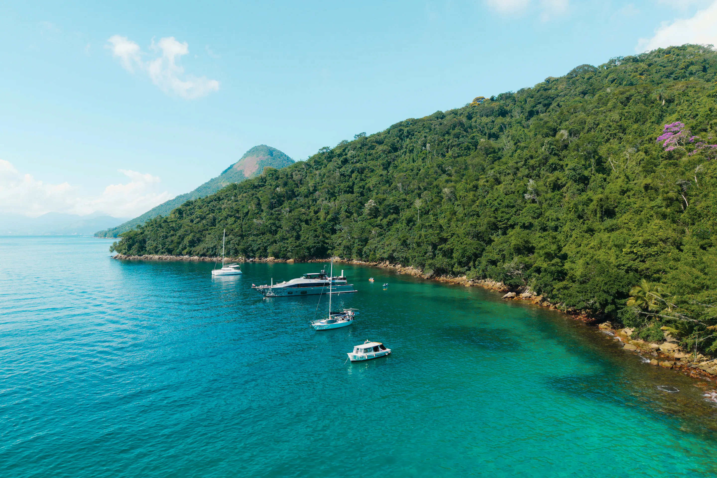 Sailboats anchor in calm turquoise water beside a densely forested green island at Ilha Grande.