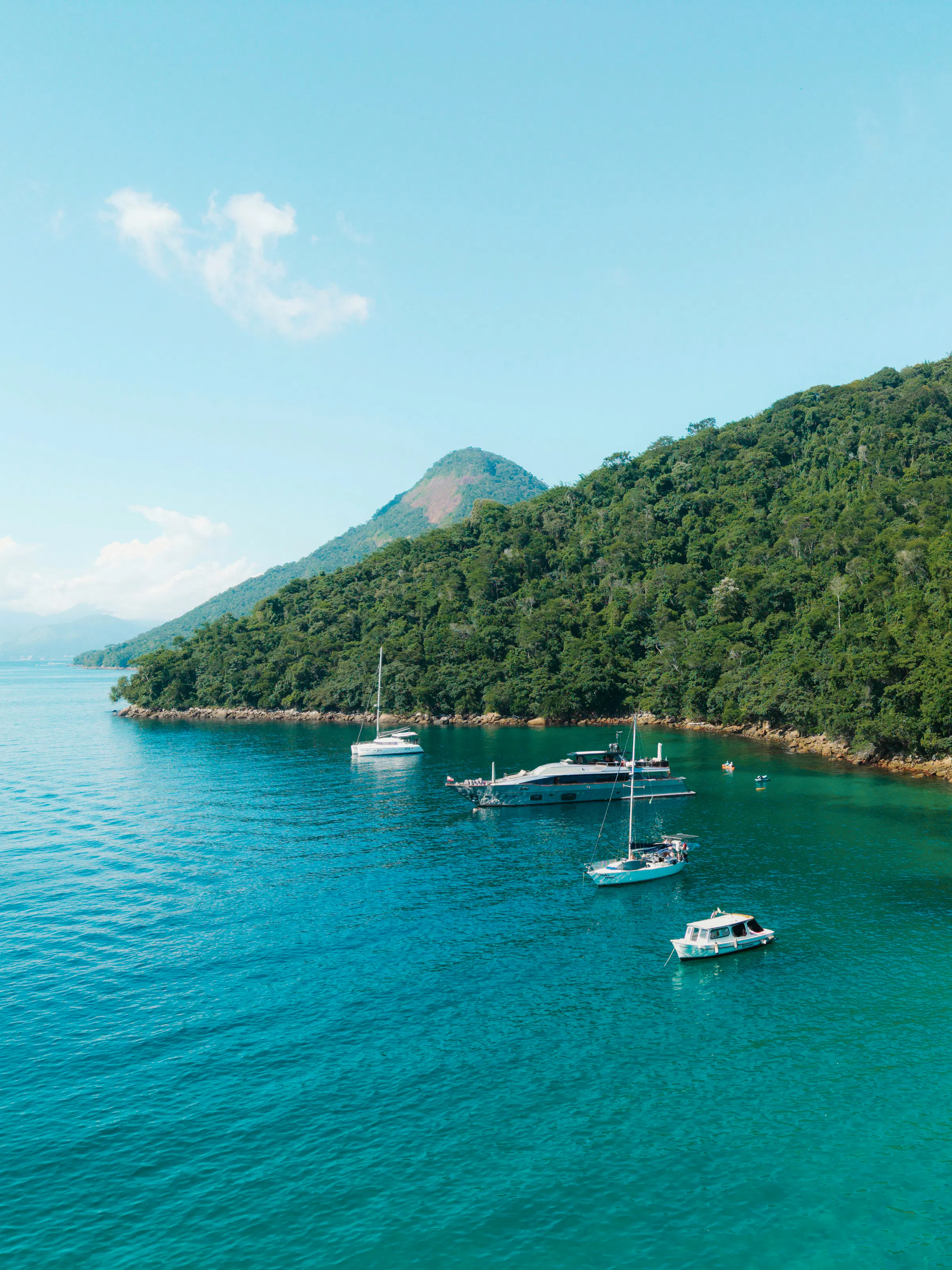 Sailboats anchor in calm turquoise water beside a densely forested green island at Ilha Grande.