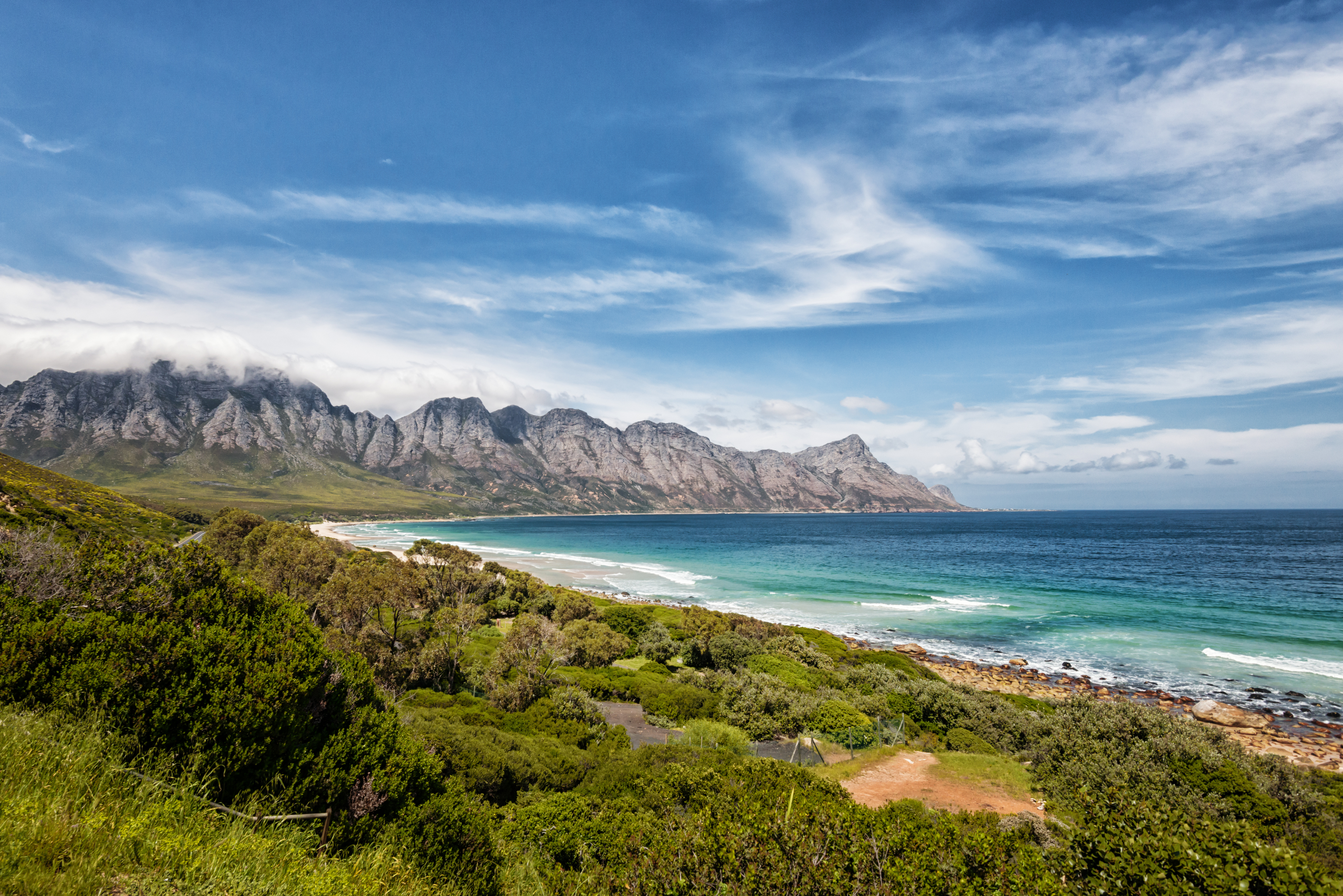Green fynbos-covered hills descend to a rocky shoreline with ocean and a pale cloudy sky beyond.