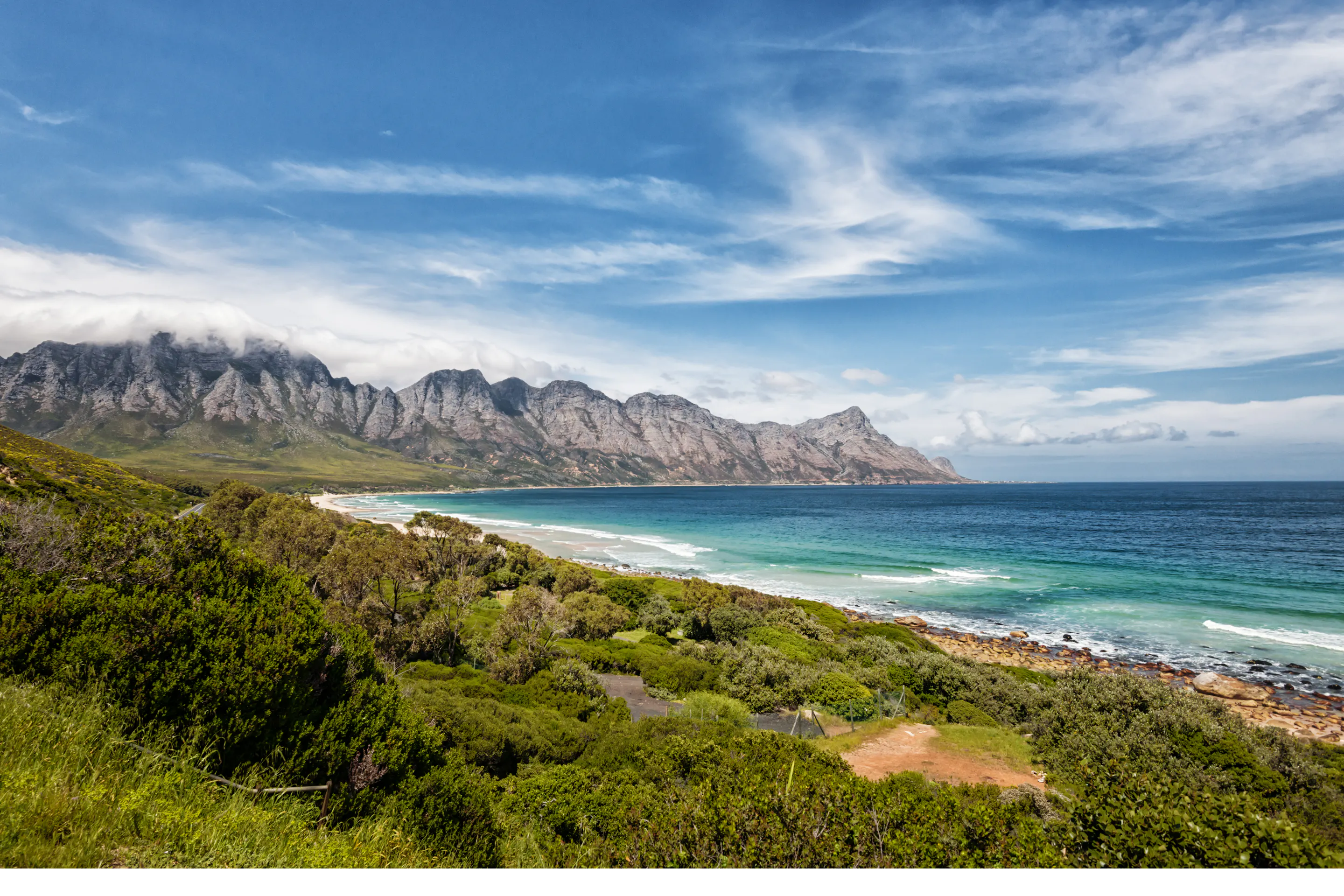 Green fynbos-covered hills descend to a rocky shoreline with ocean and a pale cloudy sky beyond.