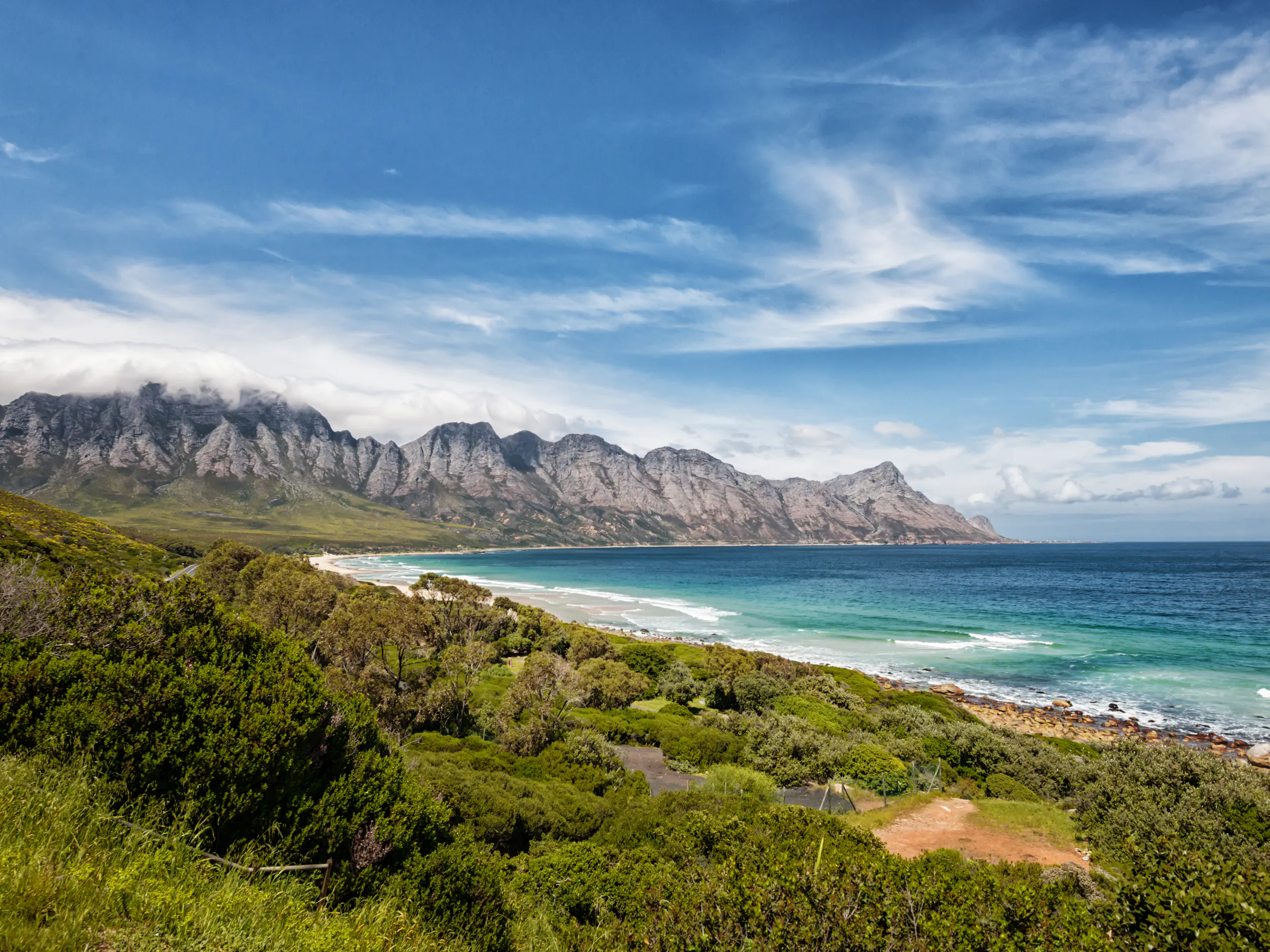 Green fynbos-covered hills descend to a rocky shoreline with ocean and a pale cloudy sky beyond.
