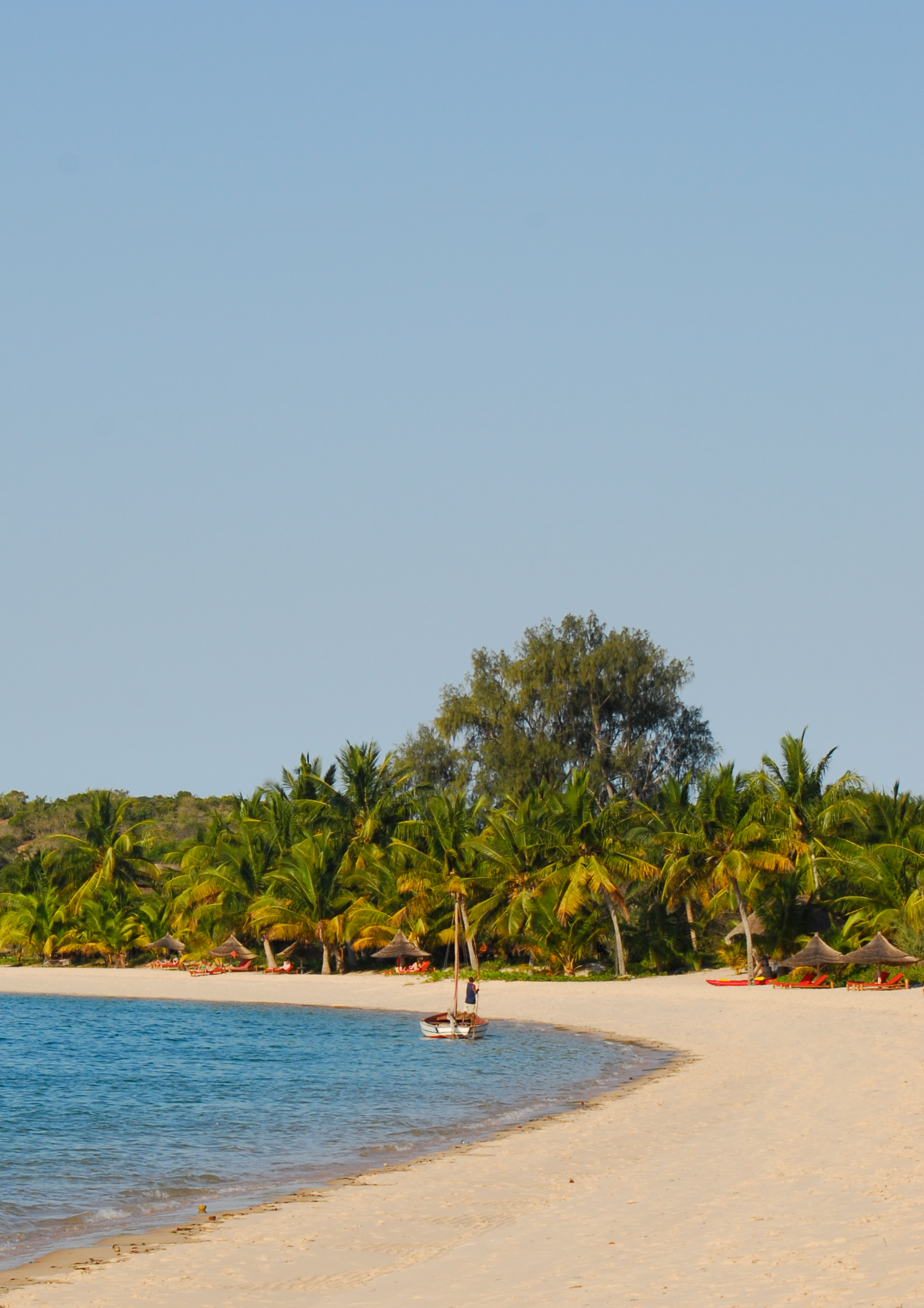A wide empty beach with pale sand meets a calm ocean beneath a broad blue sky in Mozambique.