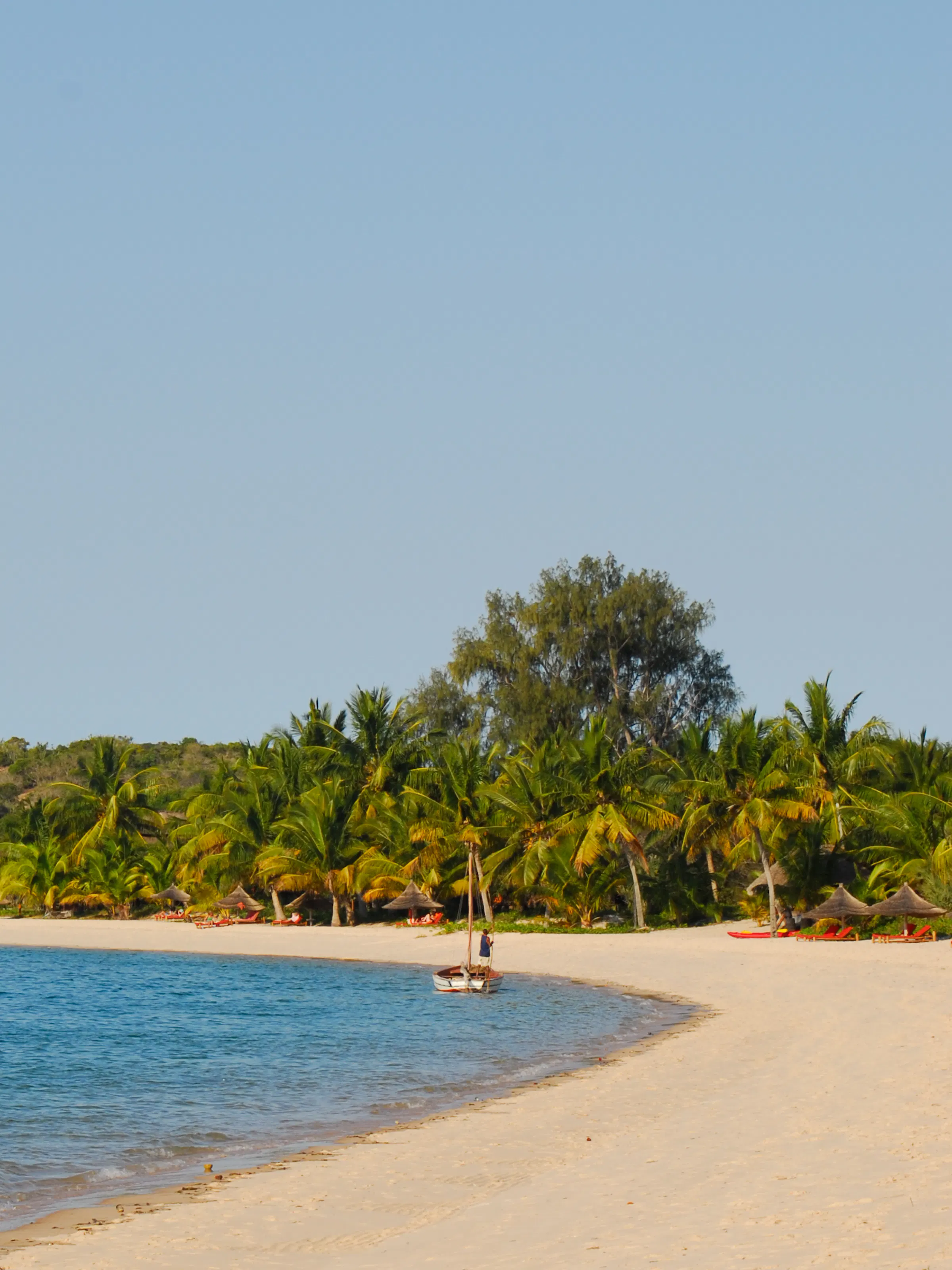 A wide empty beach with pale sand meets a calm ocean beneath a broad blue sky in Mozambique.
