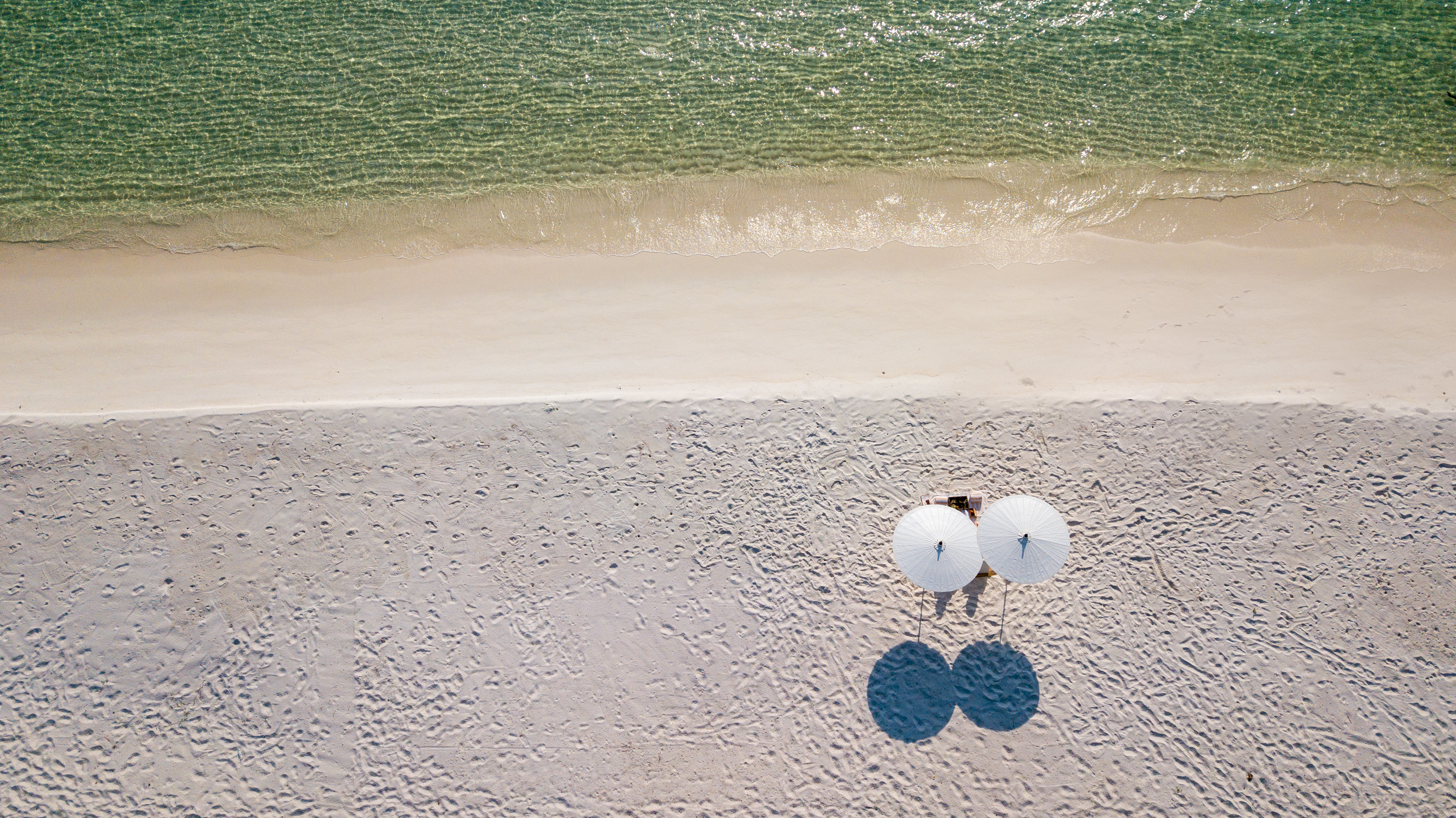 Two tiny figures stand on a remote white sand beach flanked by turquoise sea on both sides.