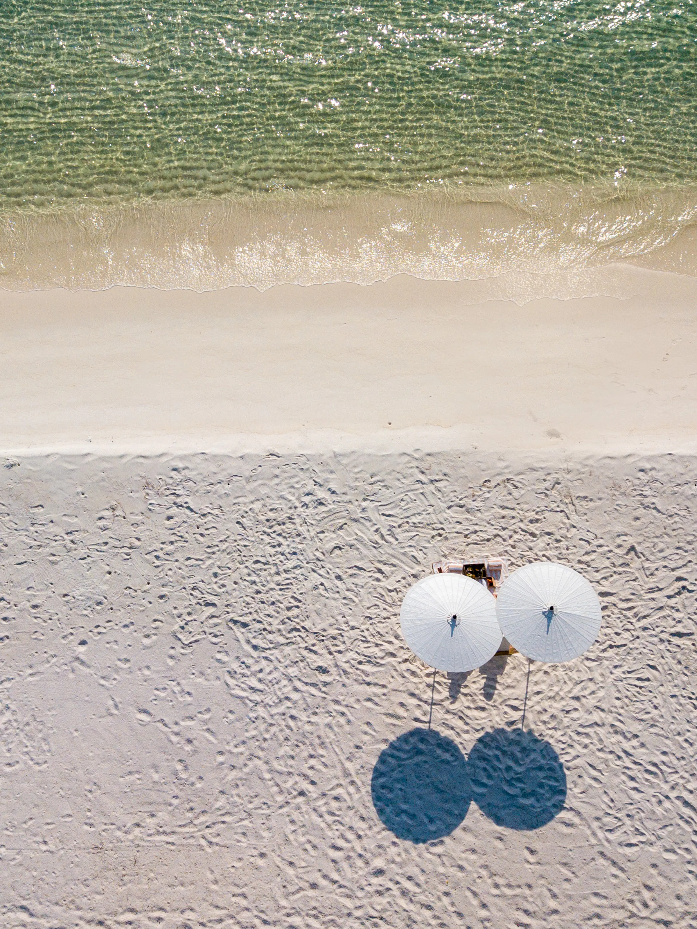 Two tiny figures stand on a remote white sand beach flanked by turquoise sea on both sides.