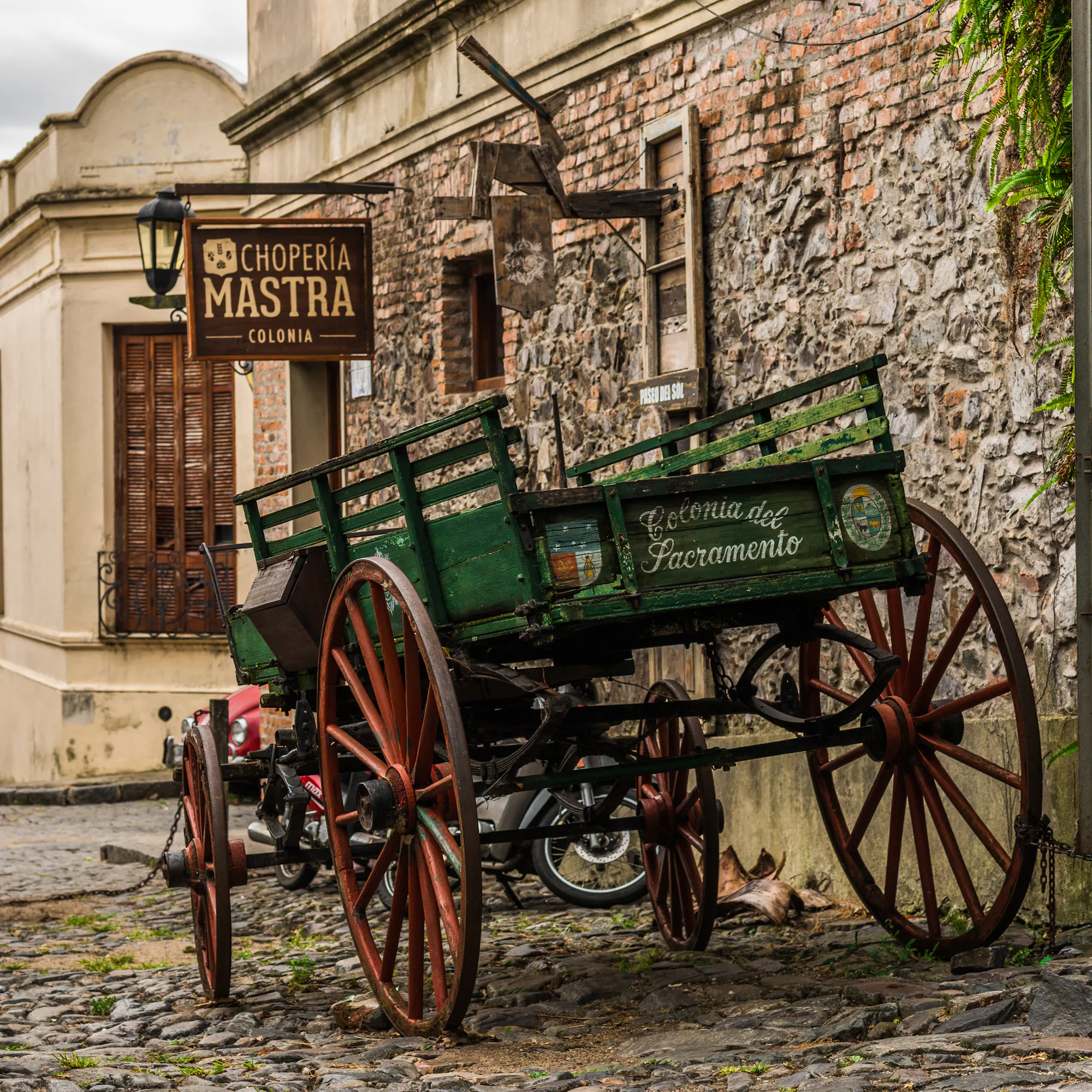 An old wooden cart sits on a stone-paved plaza in front of a colonial building in Uruguay.