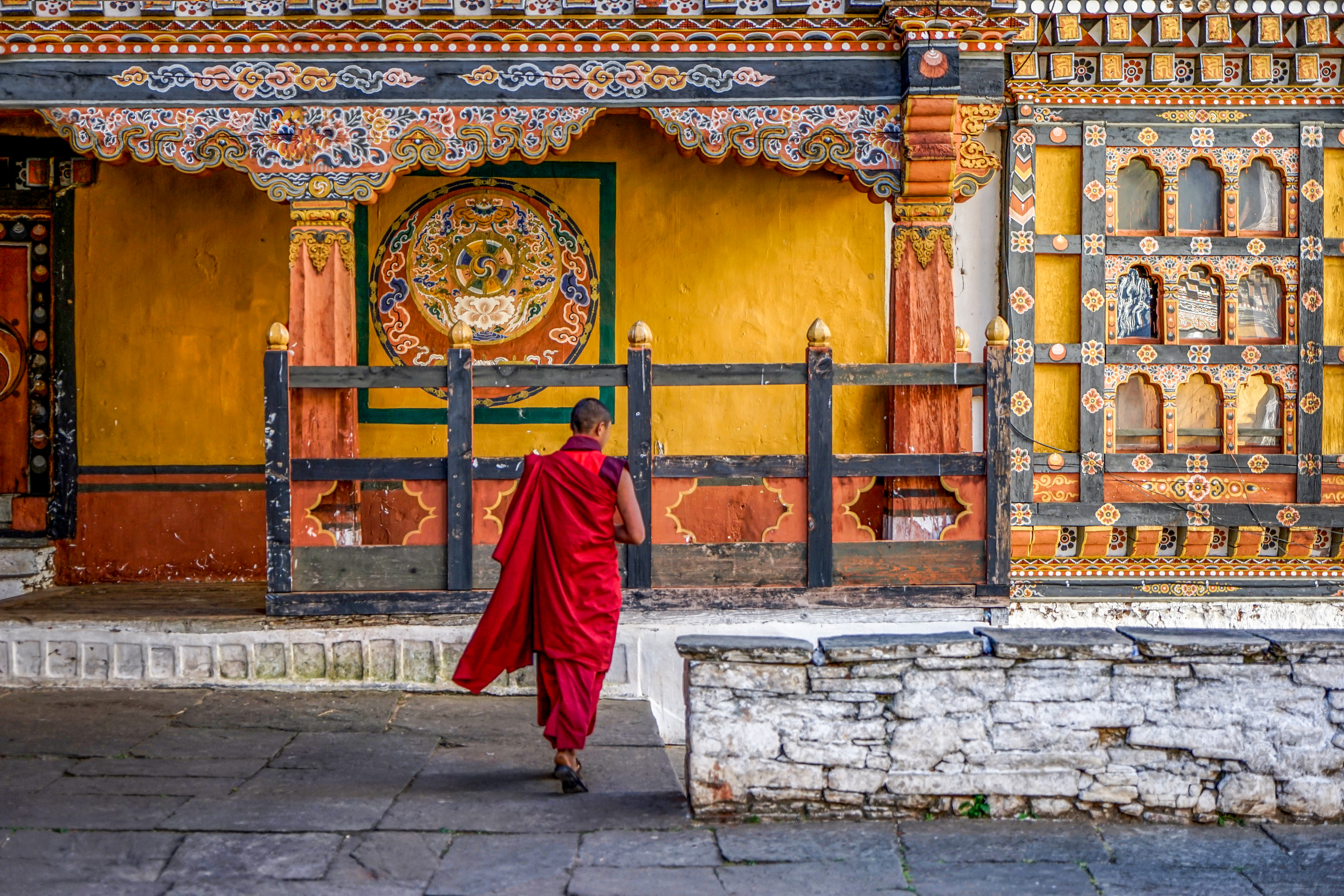 A monk in red robes walks past a richly painted yellow wall with traditional Bhutanese motifs.