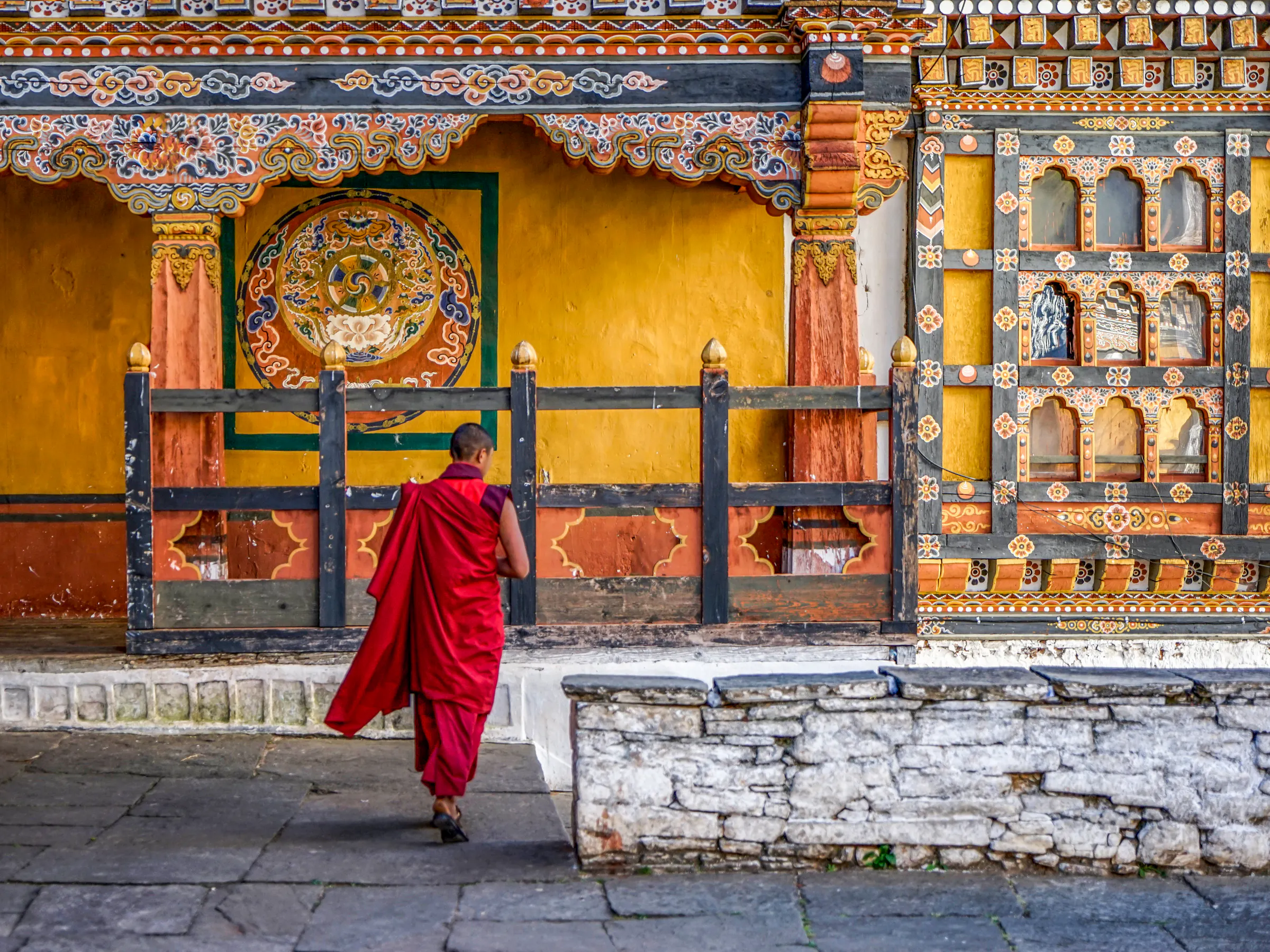 A monk in red robes walks past a richly painted yellow wall with traditional Bhutanese motifs.