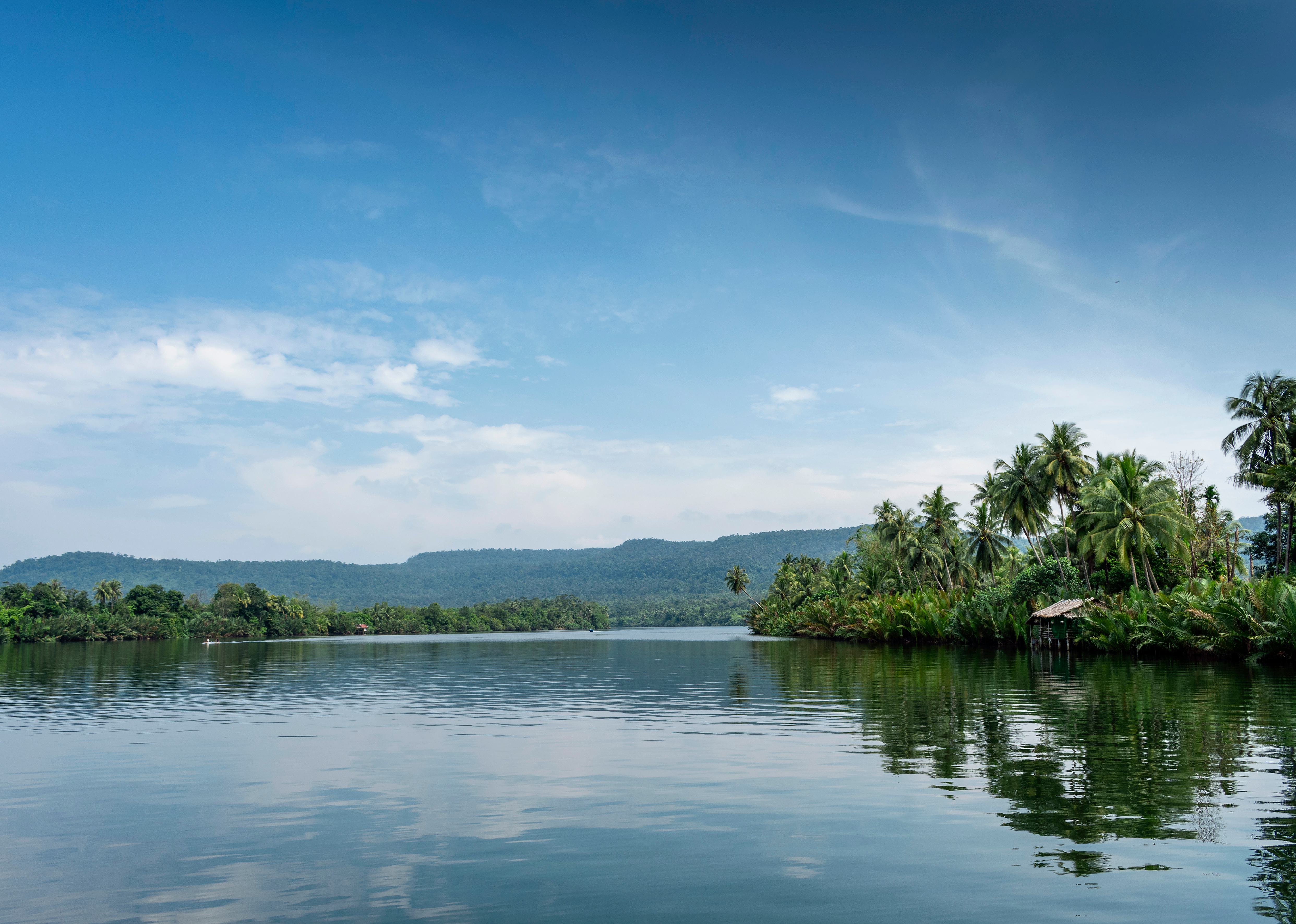 A still lake reflects green tree-covered hills under low cloud in a tranquil Cambodian landscape.