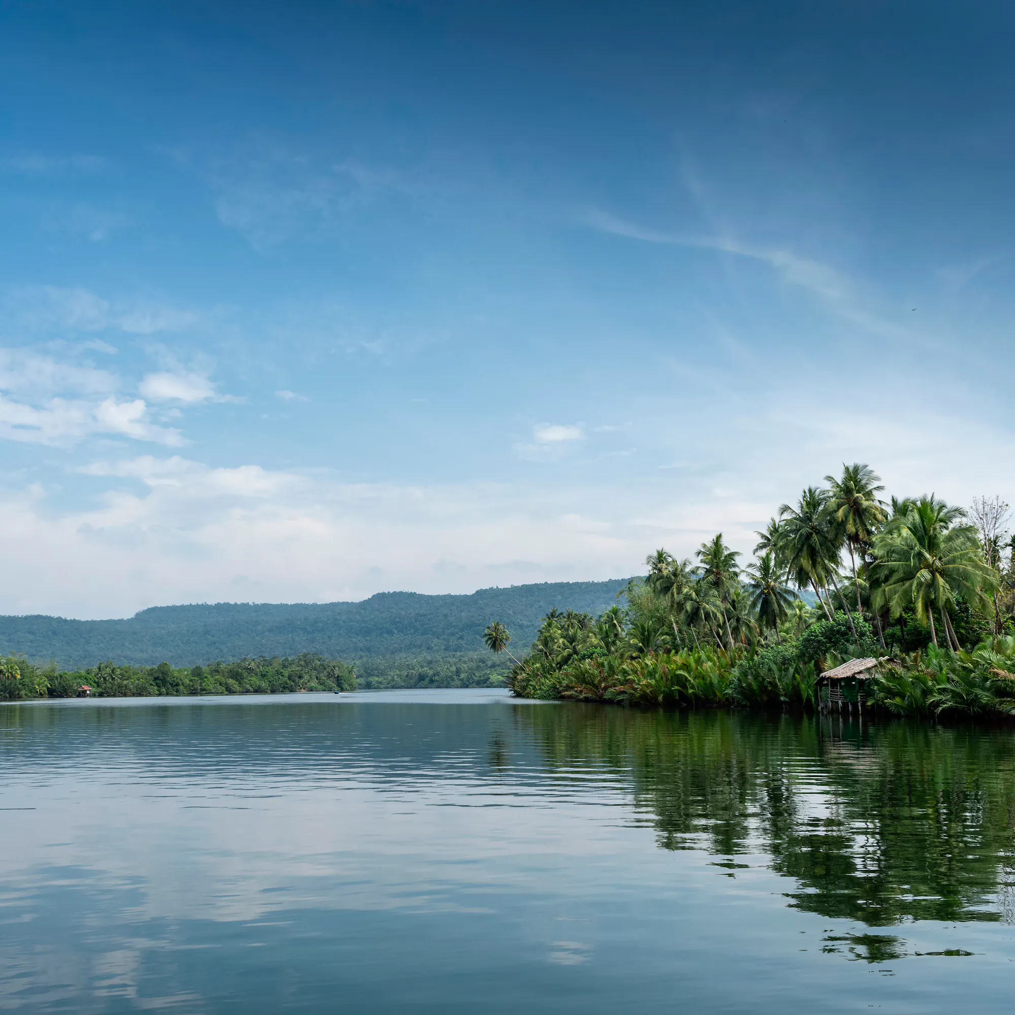 A still lake reflects green tree-covered hills under low cloud in a tranquil Cambodian landscape.