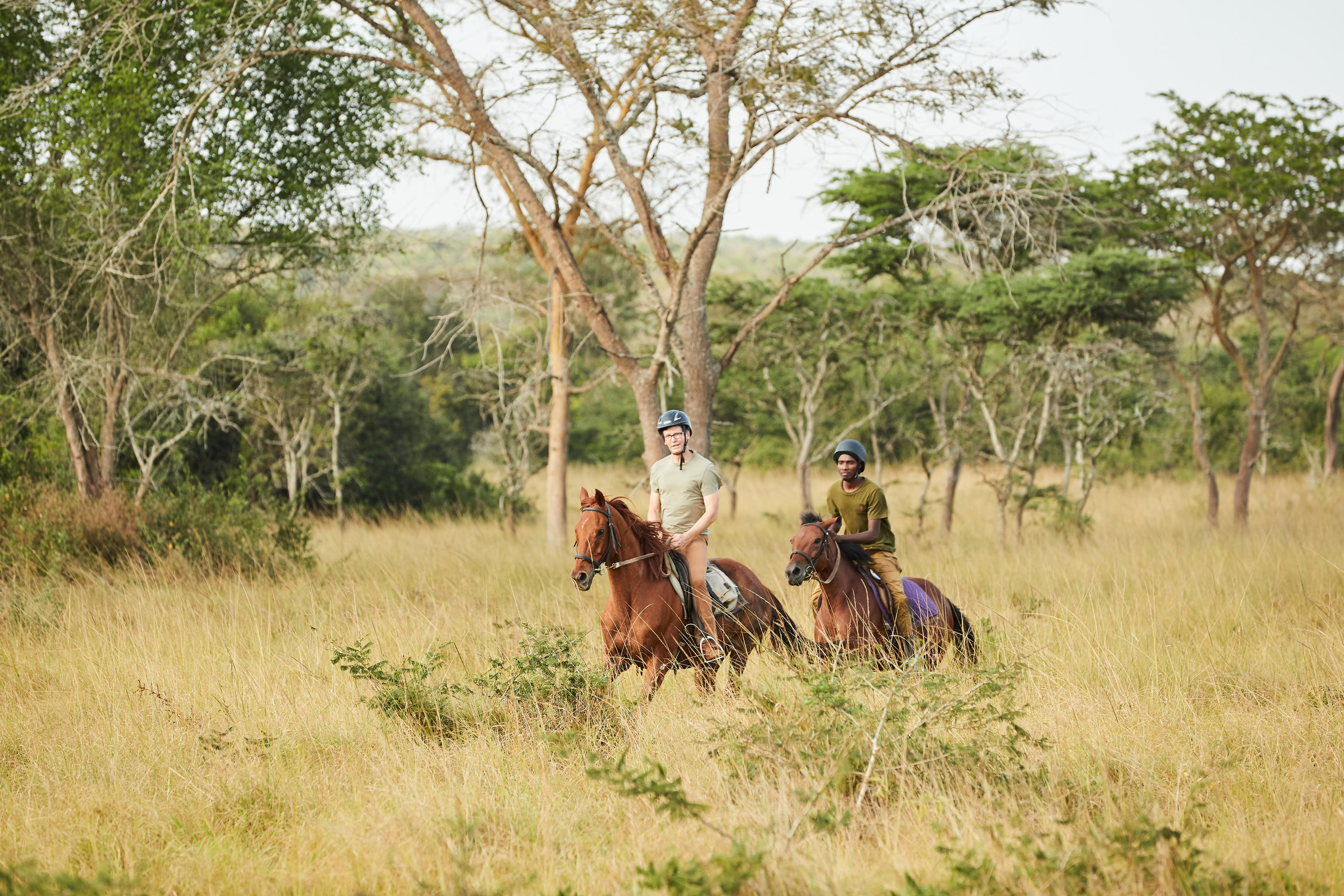 Two riders on horseback move through open savanna woodland with acacia trees in Uganda.