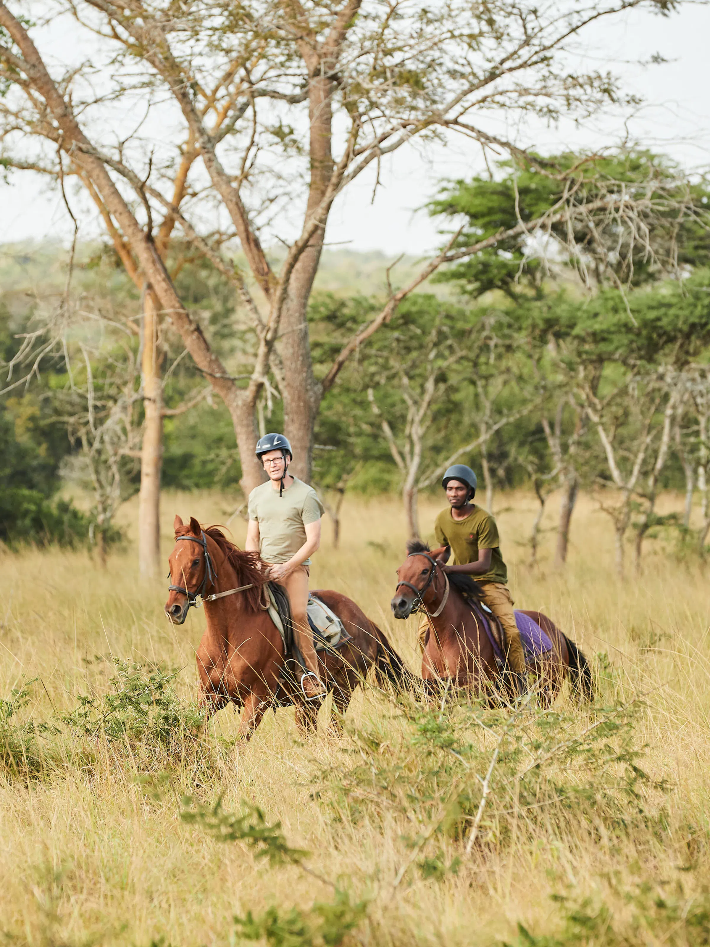 Two riders on horseback move through open savanna woodland with acacia trees in Uganda.