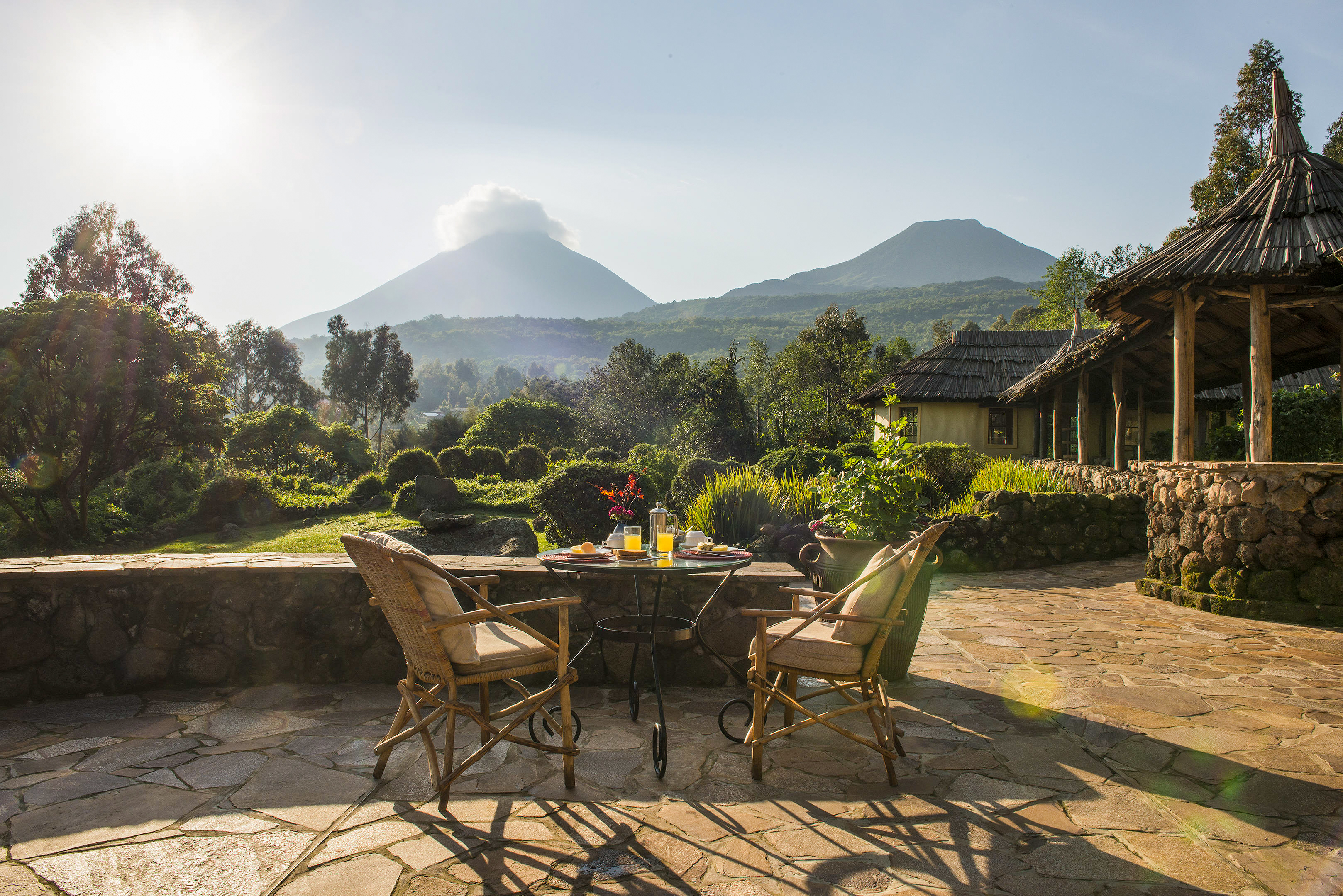 A lodge terrace with wicker chairs overlooks a green garden with a Virunga volcano beyond.