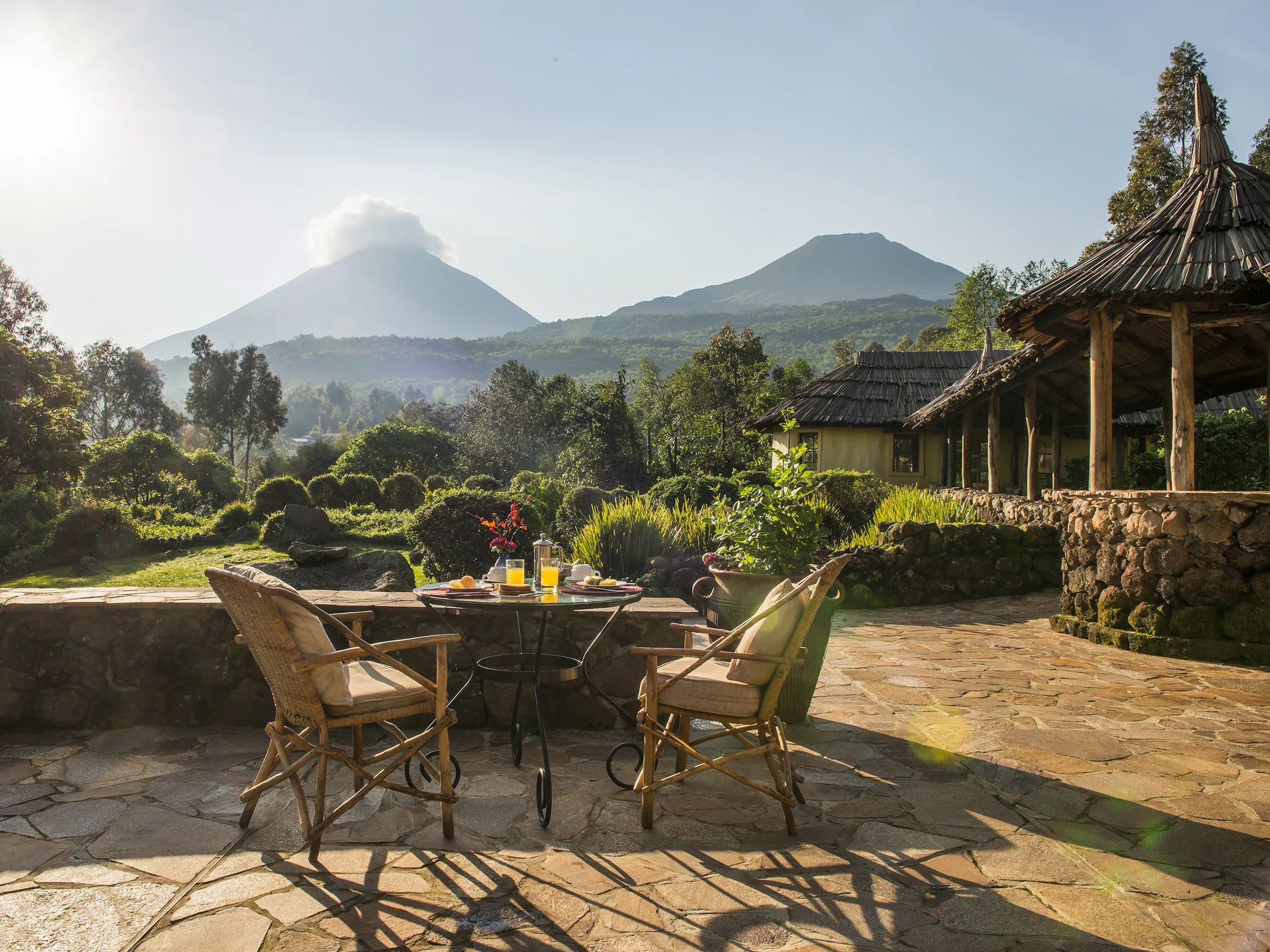 A lodge terrace with wicker chairs overlooks a green garden with a Virunga volcano beyond.