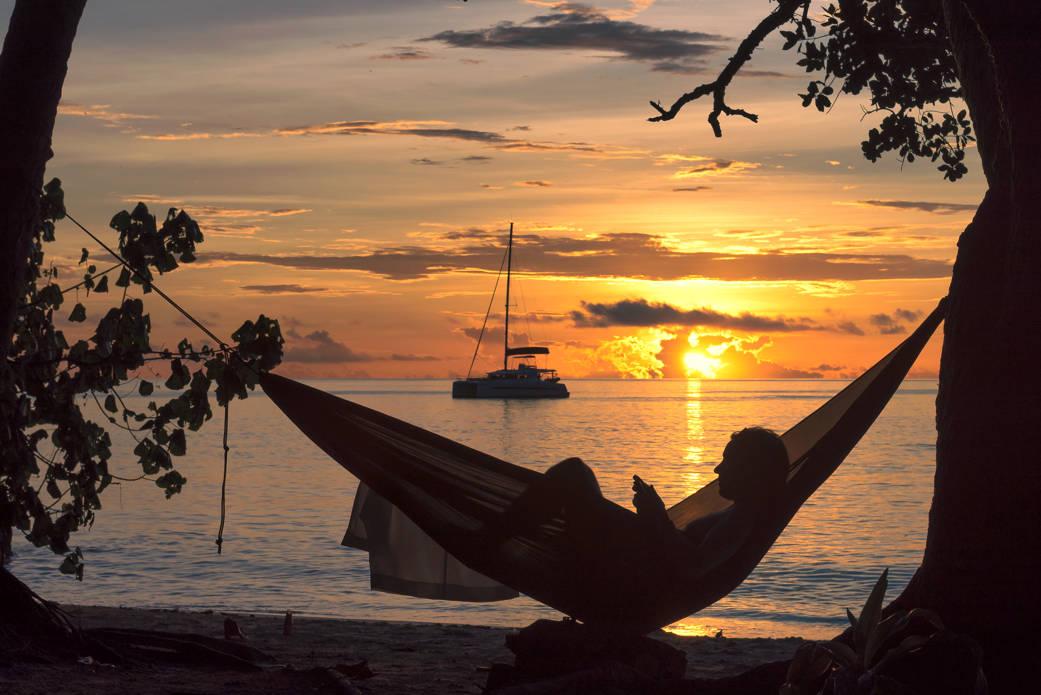 A person reclines in a hammock strung between palm trees above a beach at golden sunset.
