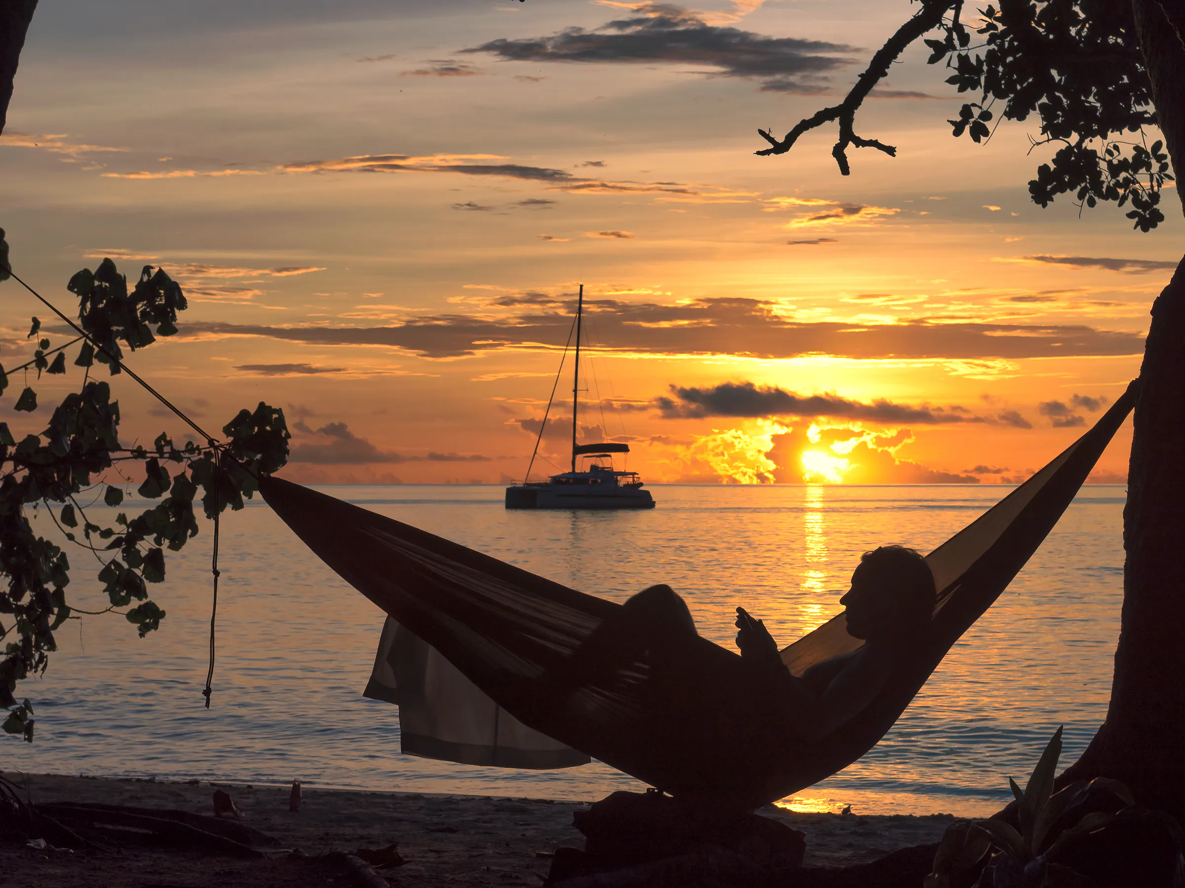A person reclines in a hammock strung between palm trees above a beach at golden sunset.