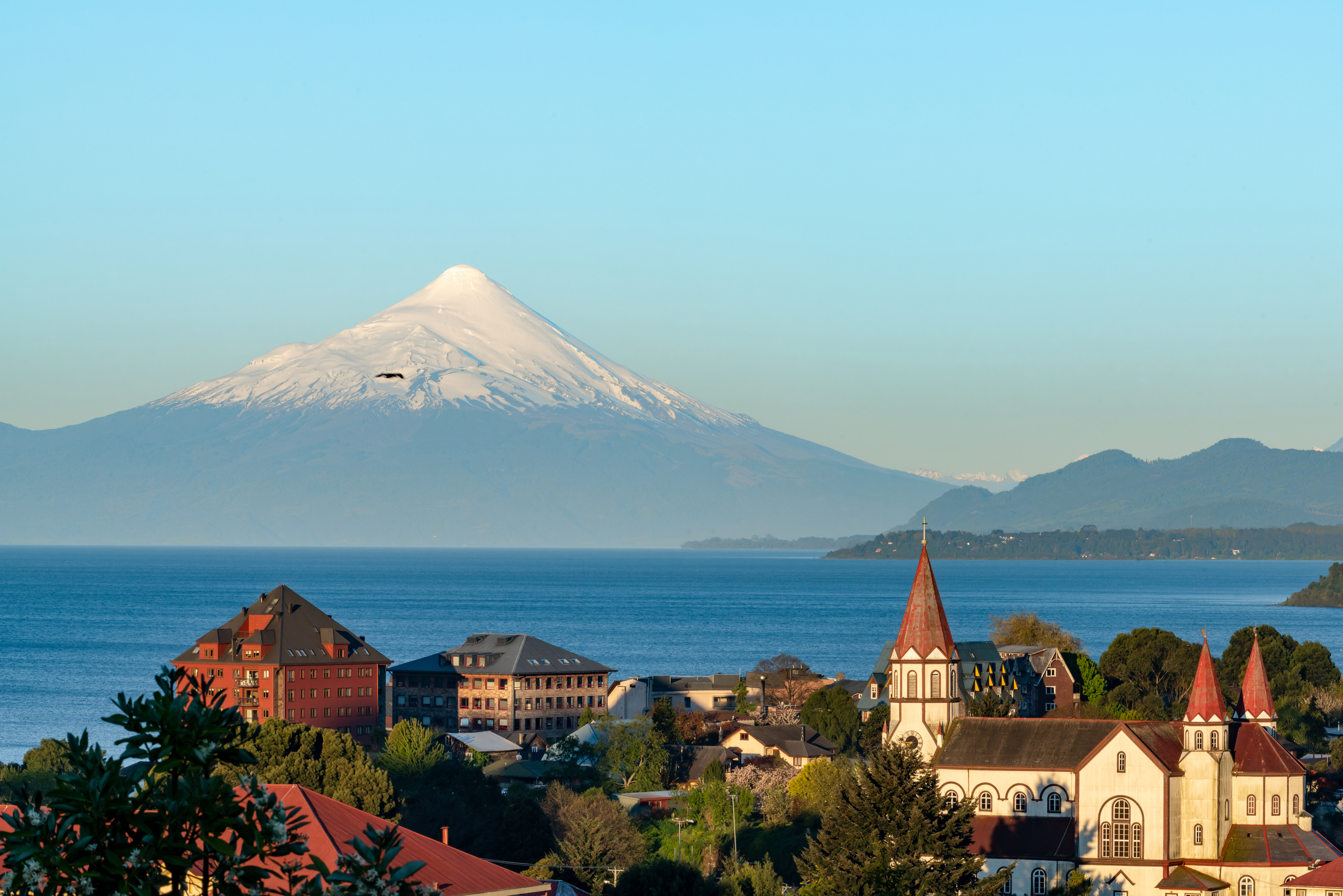 A snow-capped volcano rises behind a European-style church and colorful lakeside town in Chile.