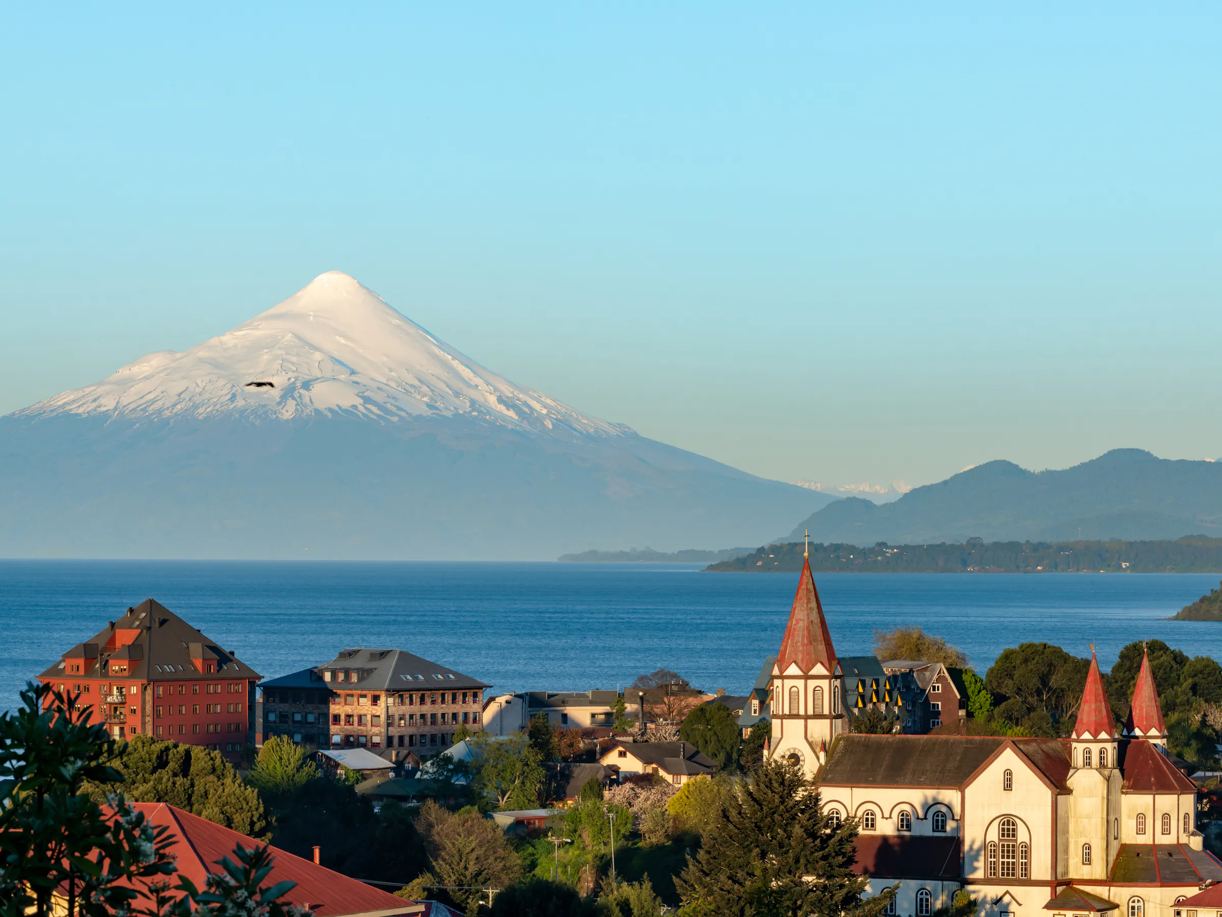 A snow-capped volcano rises behind a European-style church and colorful lakeside town in Chile.