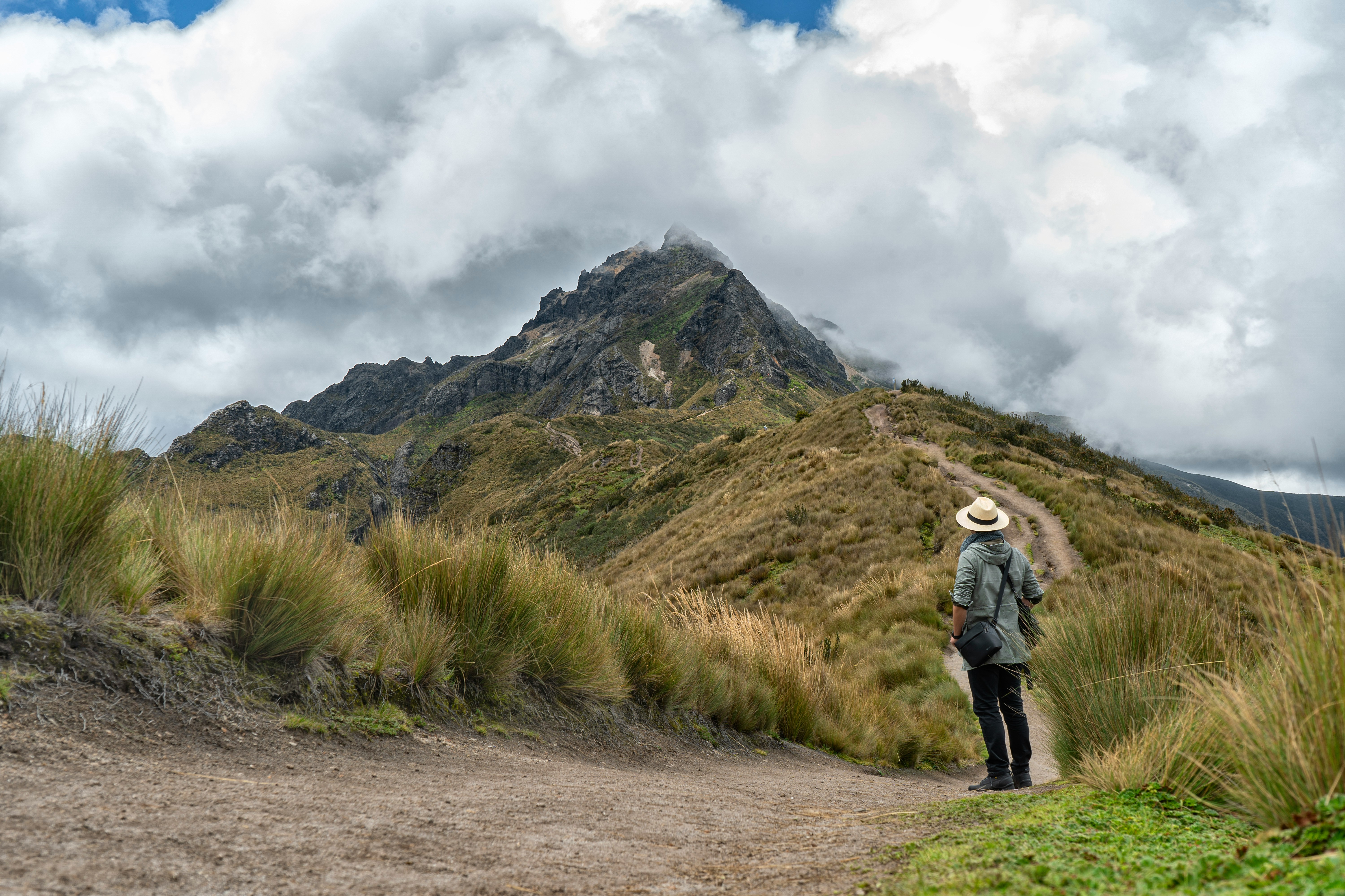A hiker stands on a rocky ridge overlooking layered green Andean peaks under a cloudy sky.