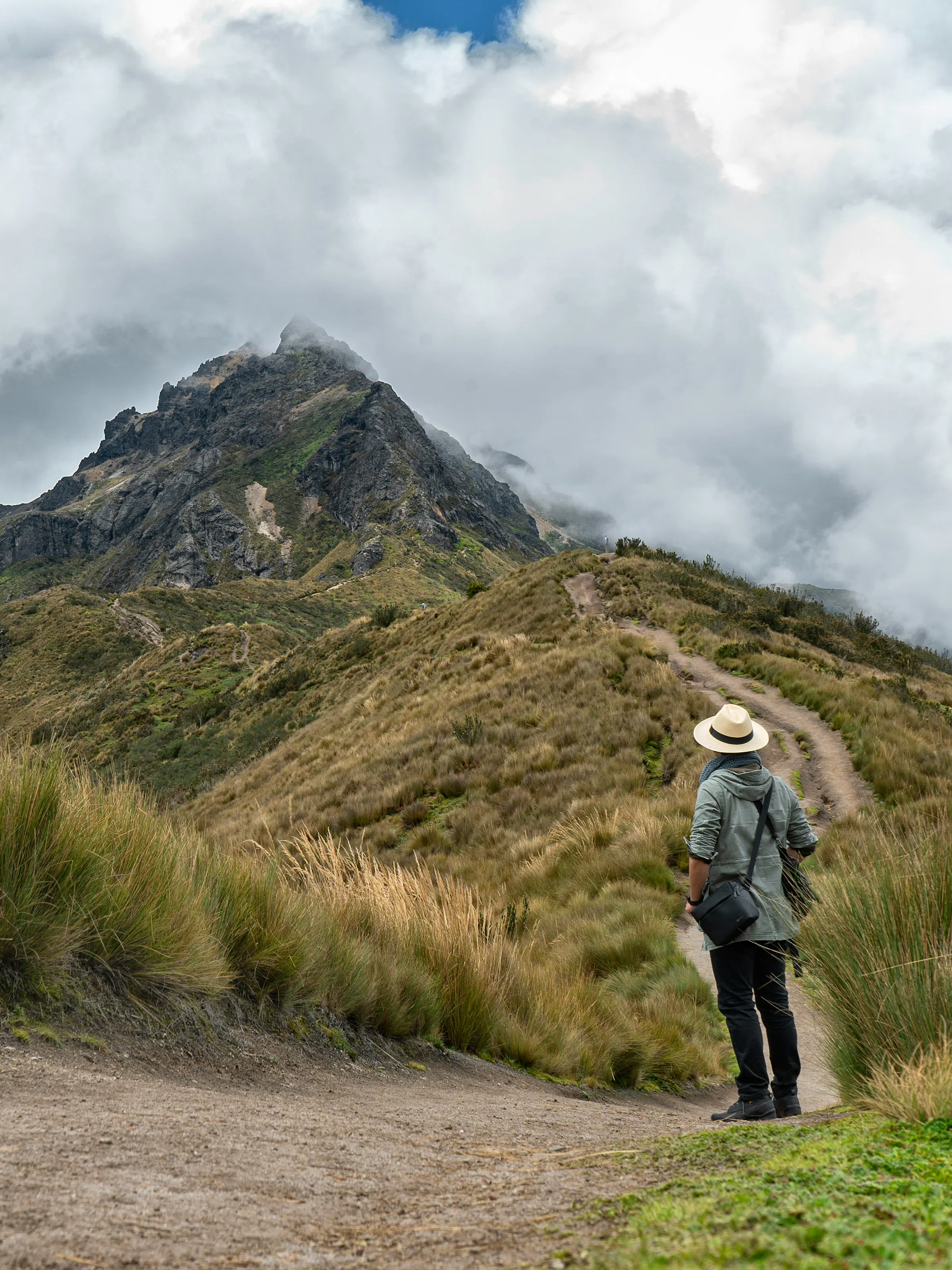 A hiker stands on a rocky ridge overlooking layered green Andean peaks under a cloudy sky.