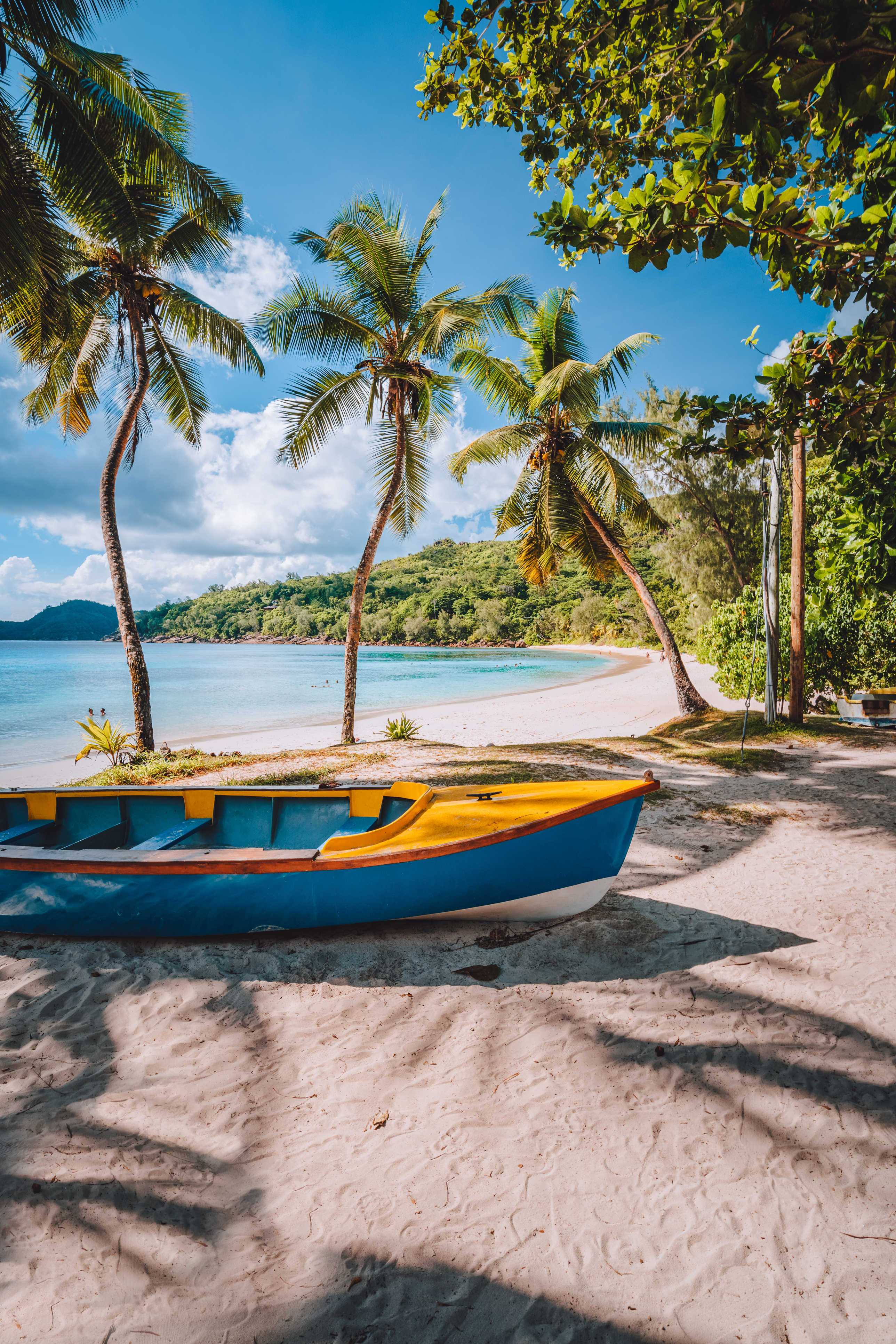 Blue wooden boats rest on a sandy beach edged with palm trees under a partly cloudy sky in Mahé.