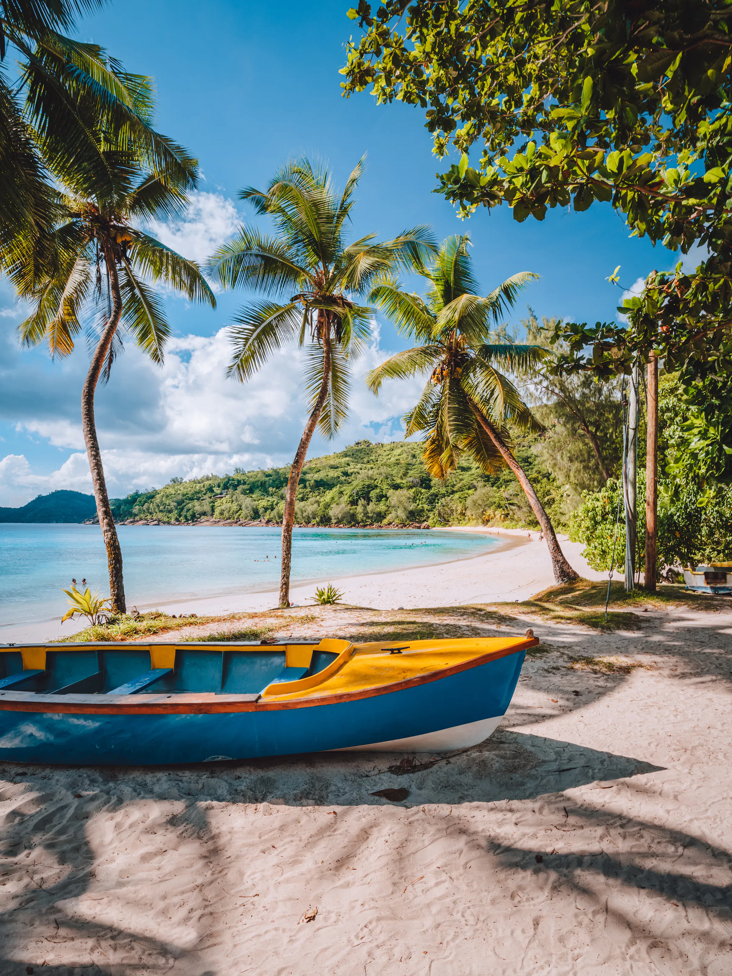 Blue wooden boats rest on a sandy beach edged with palm trees under a partly cloudy sky in Mahé.
