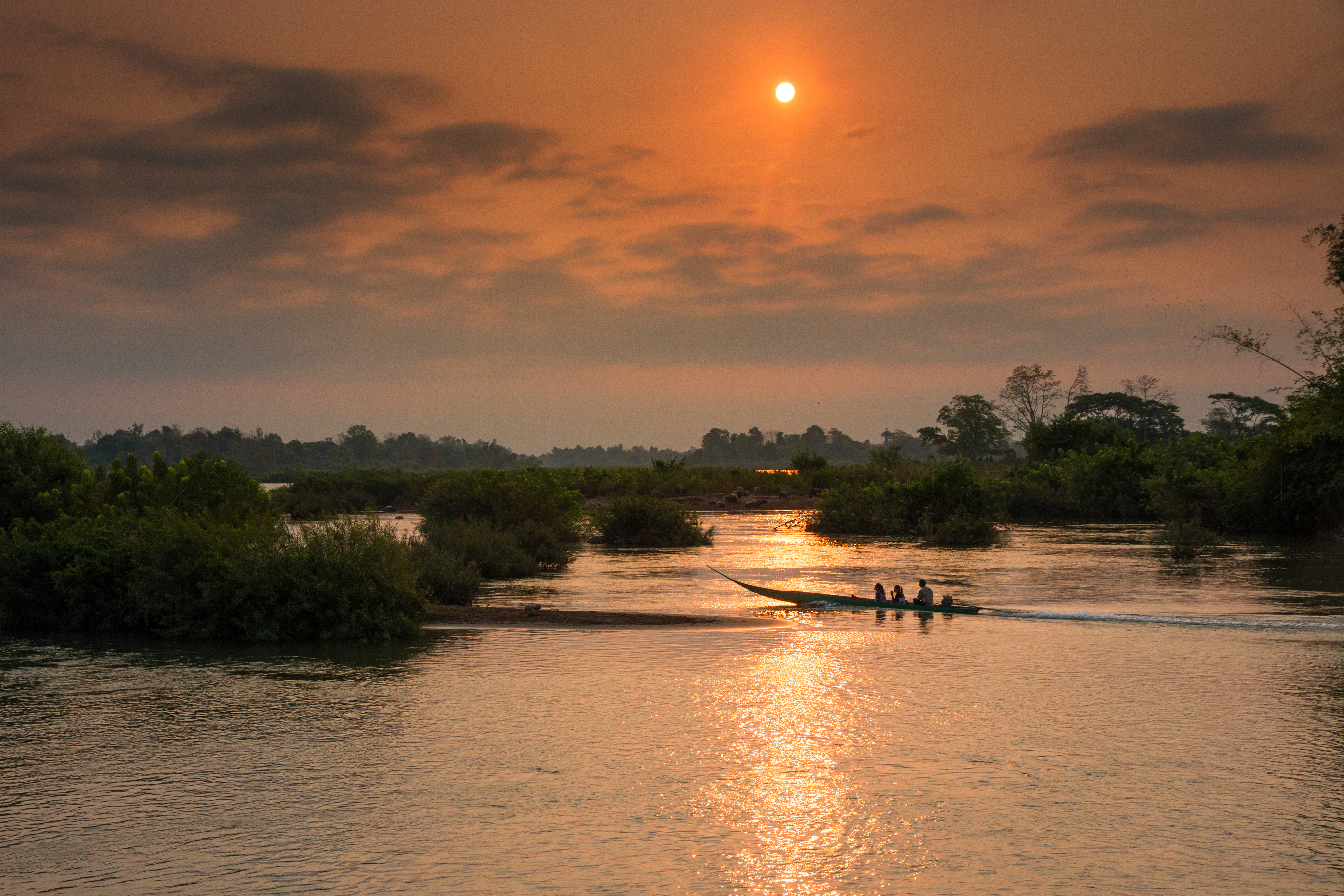 A small wooden boat moves along a wide river at sunset, orange light reflecting on the water.