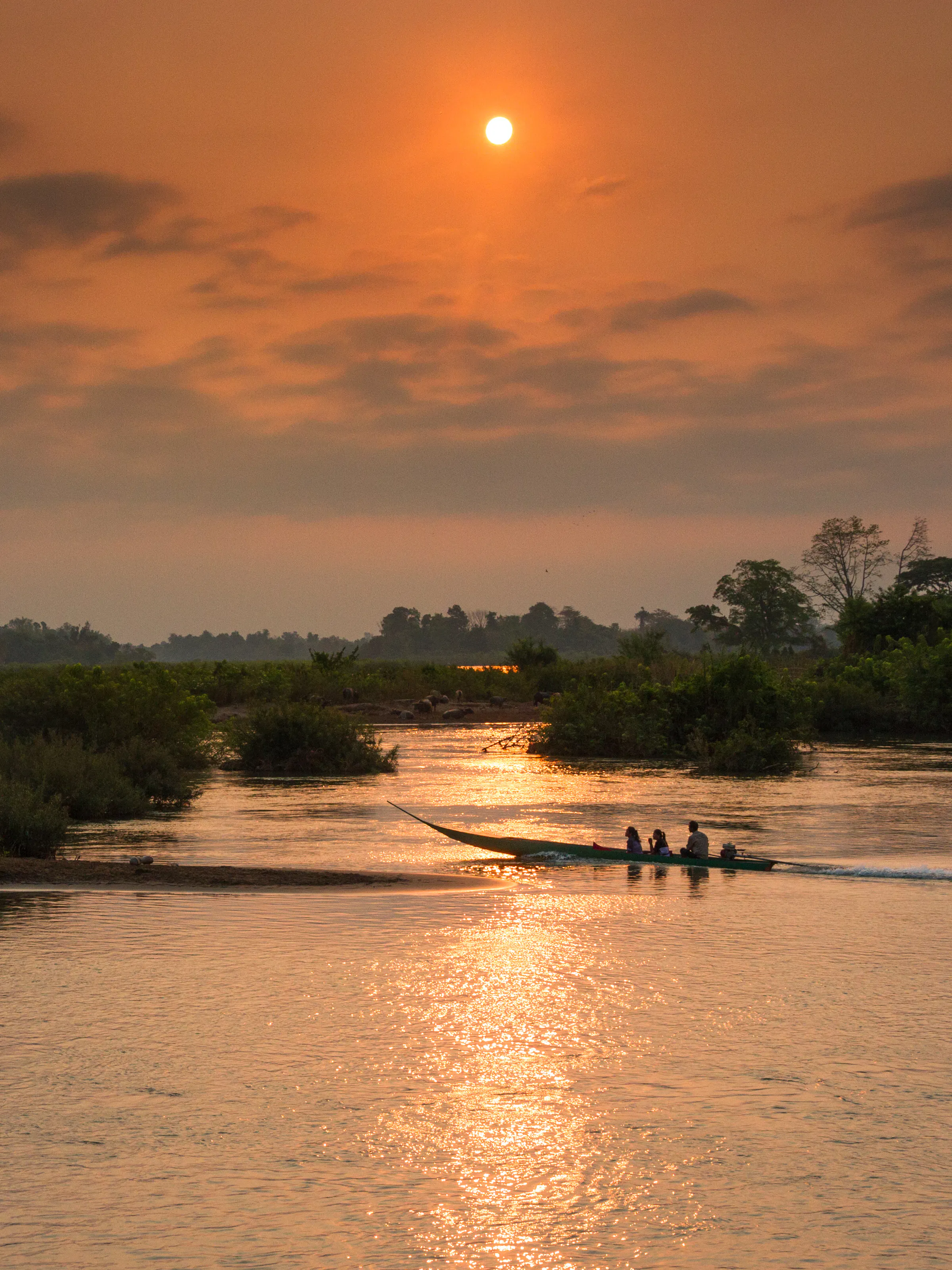 A small wooden boat moves along a wide river at sunset, orange light reflecting on the water.