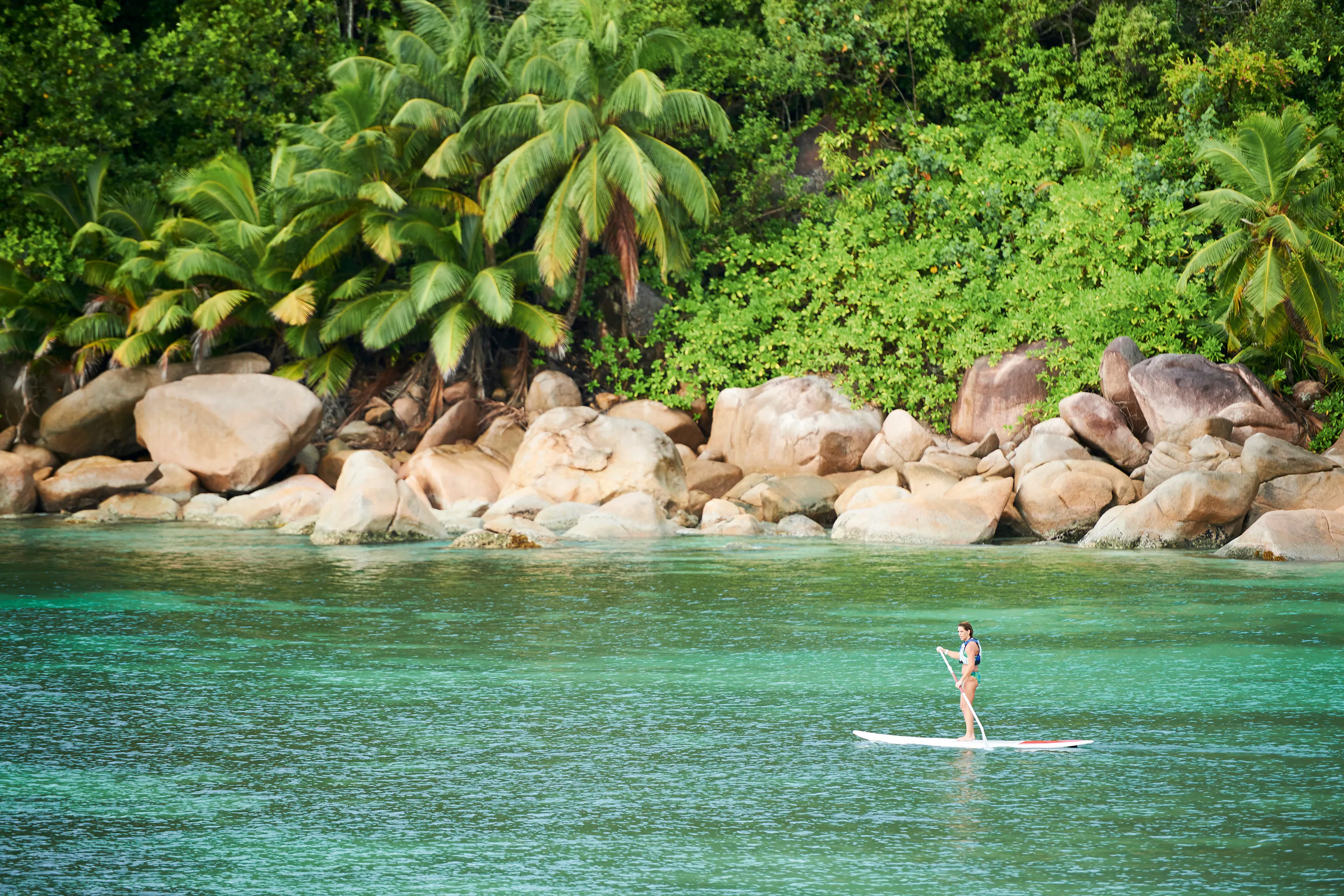A person stand-up paddleboards on clear shallow water beside a rocky granite shoreline in Seychelles.