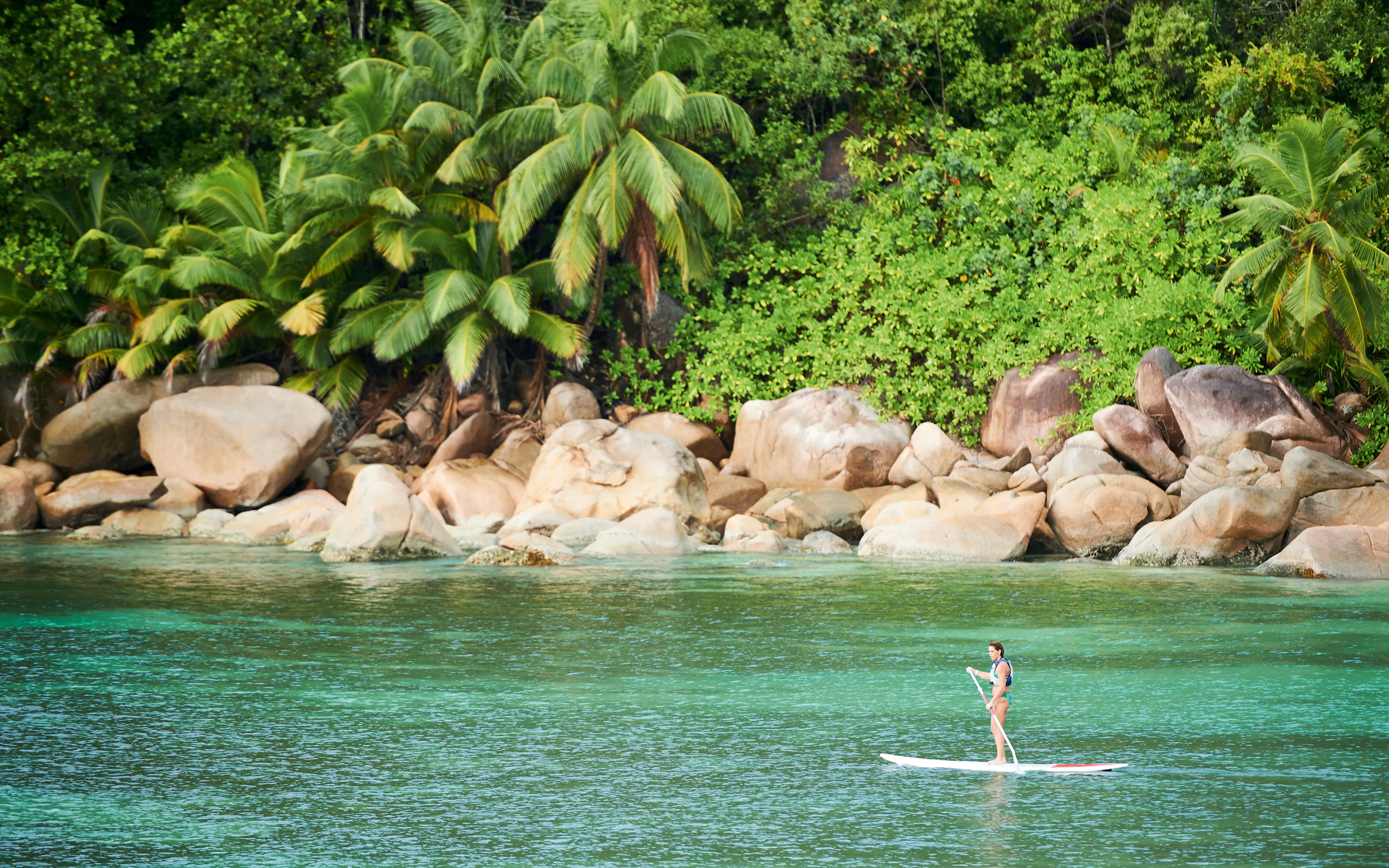 A person stand-up paddleboards on clear shallow water beside a rocky granite shoreline in Seychelles.
