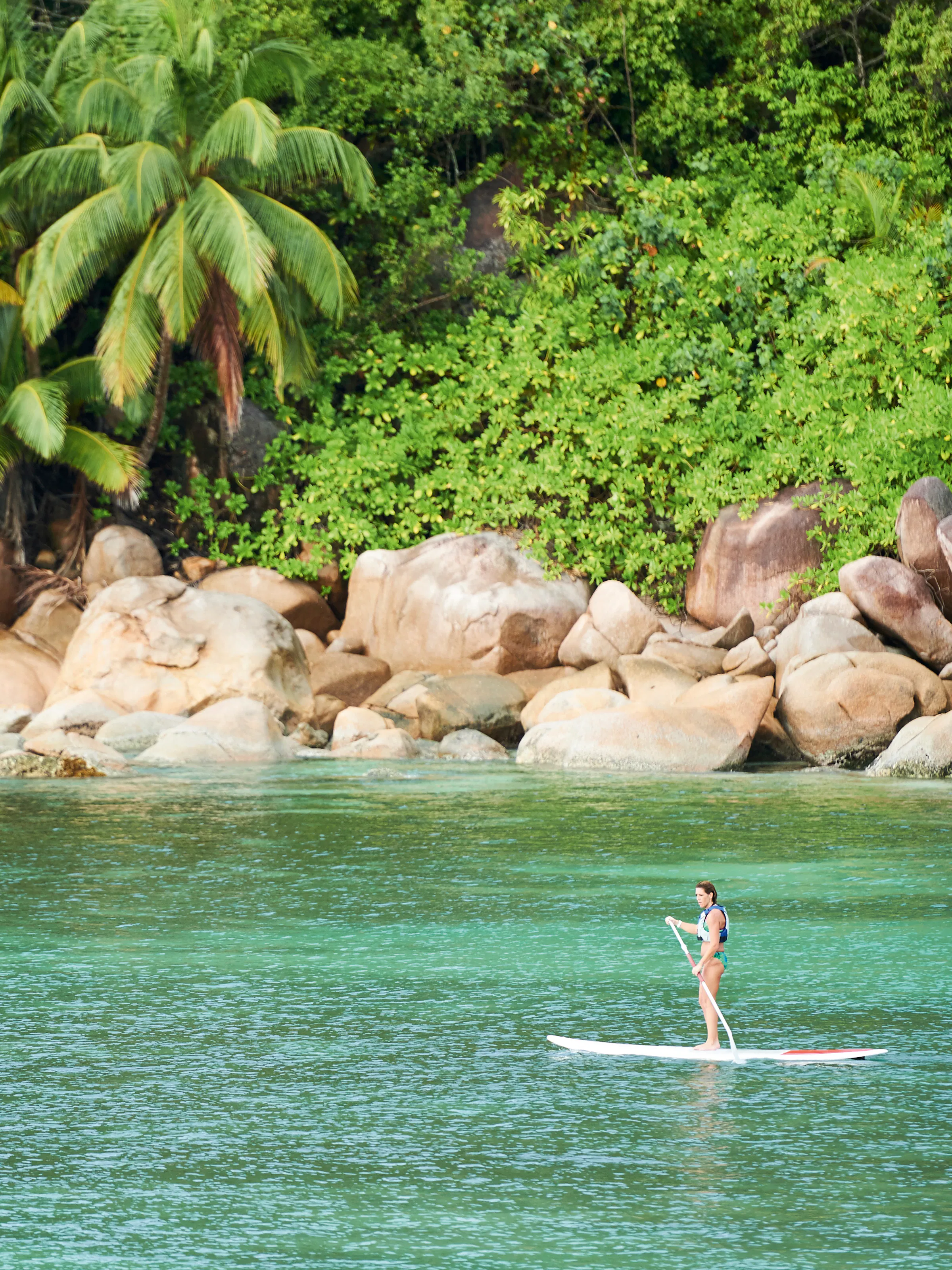 A person stand-up paddleboards on clear shallow water beside a rocky granite shoreline in Seychelles.