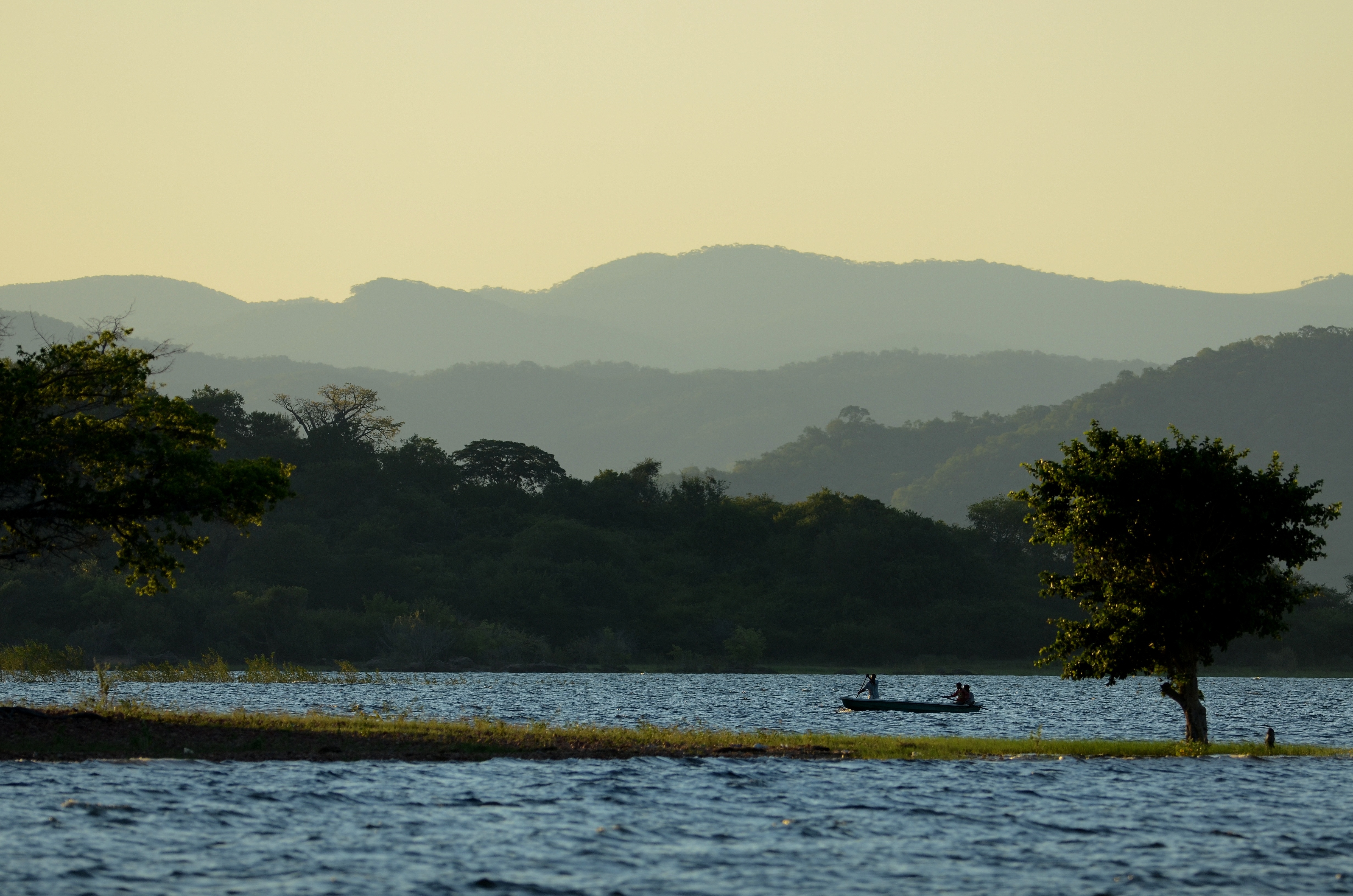 A small boat moves along a wide calm lake flanked by green hills under soft overcast light.