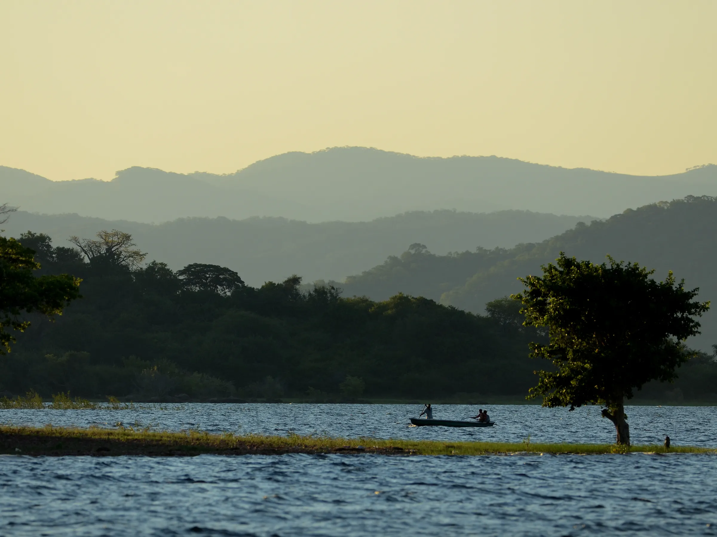 A small boat moves along a wide calm lake flanked by green hills under soft overcast light.
