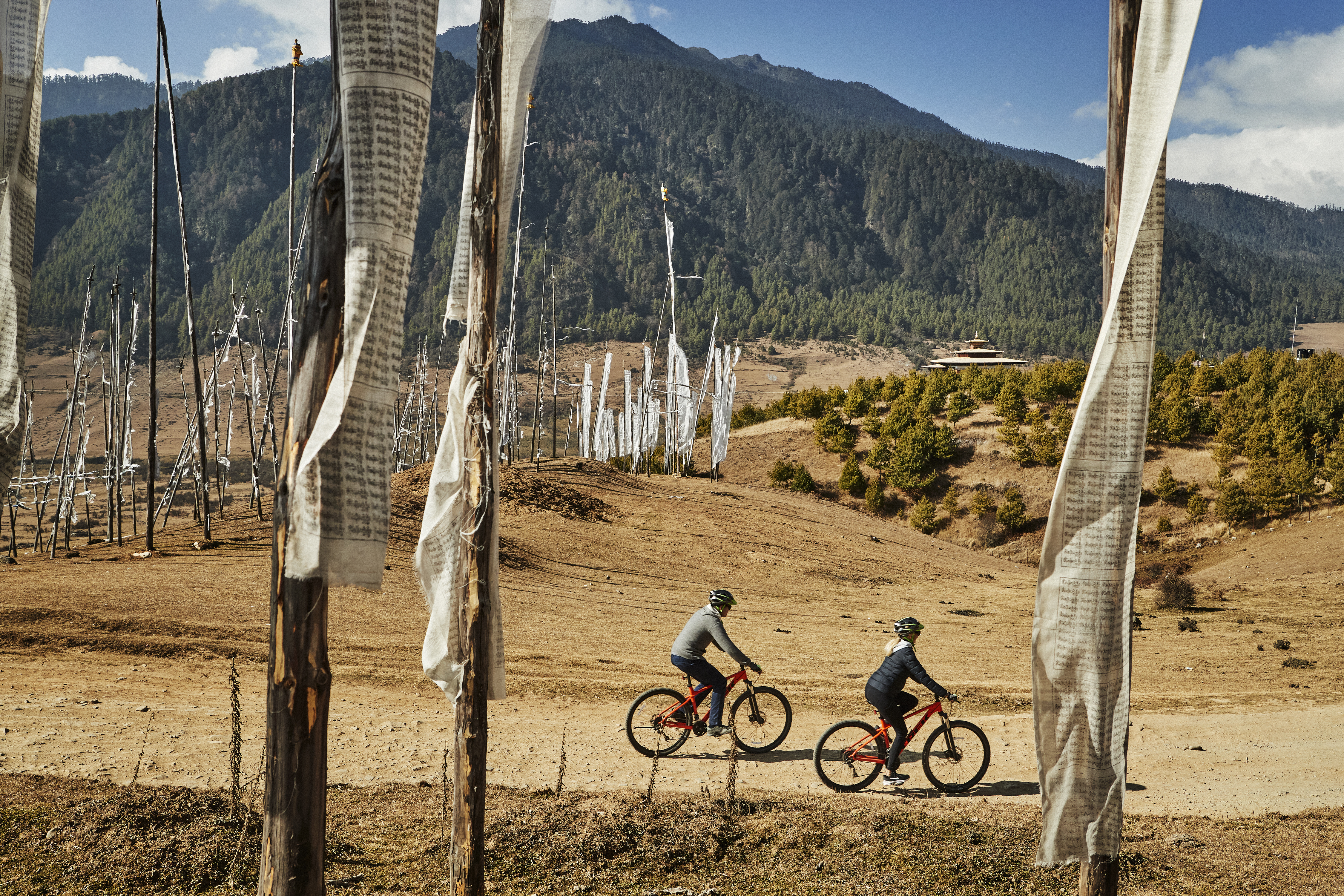 Two cyclists ride a dirt trail lined with colorful prayer flags across a high-altitude Bhutanese valley.