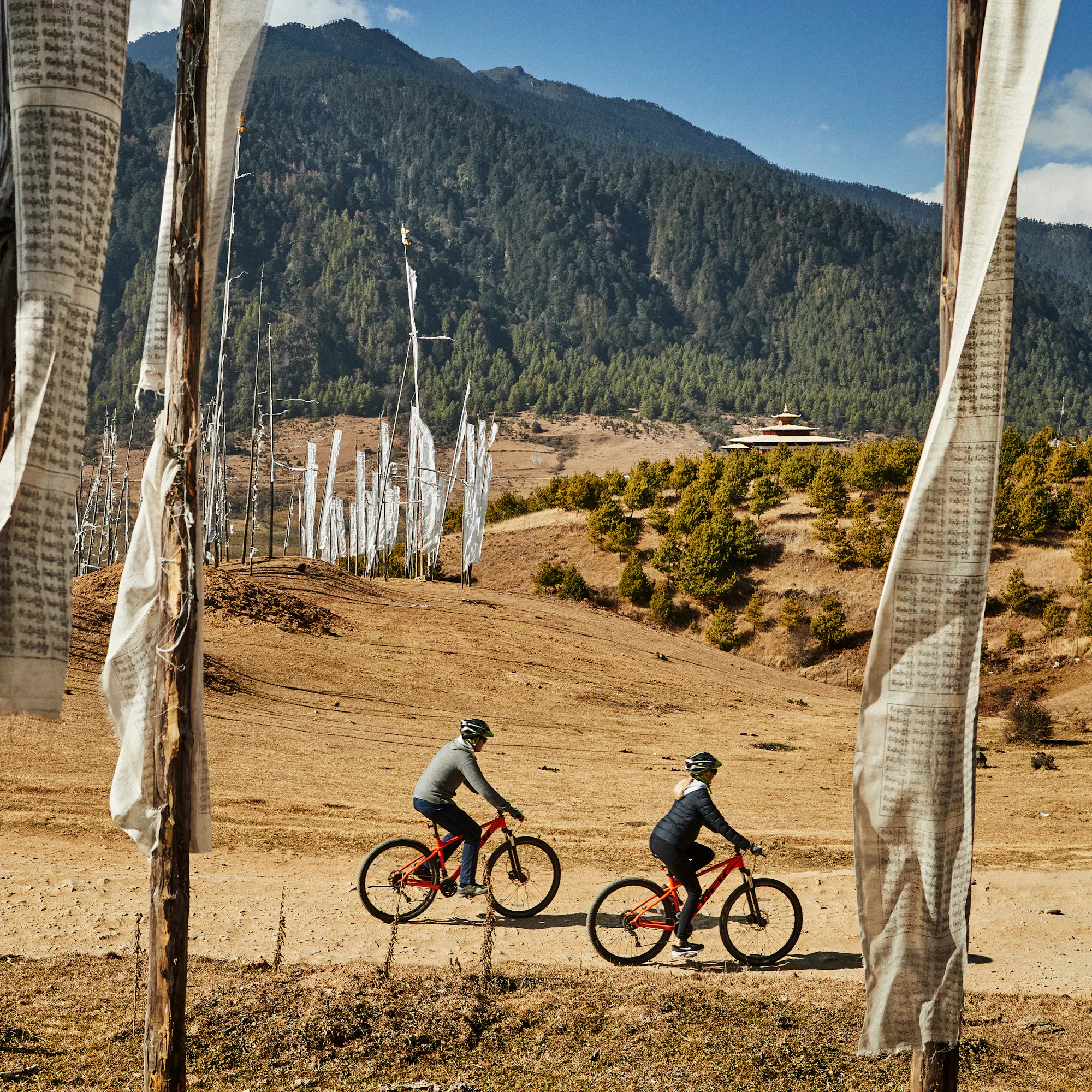 Two cyclists ride a dirt trail lined with colorful prayer flags across a high-altitude Bhutanese valley.