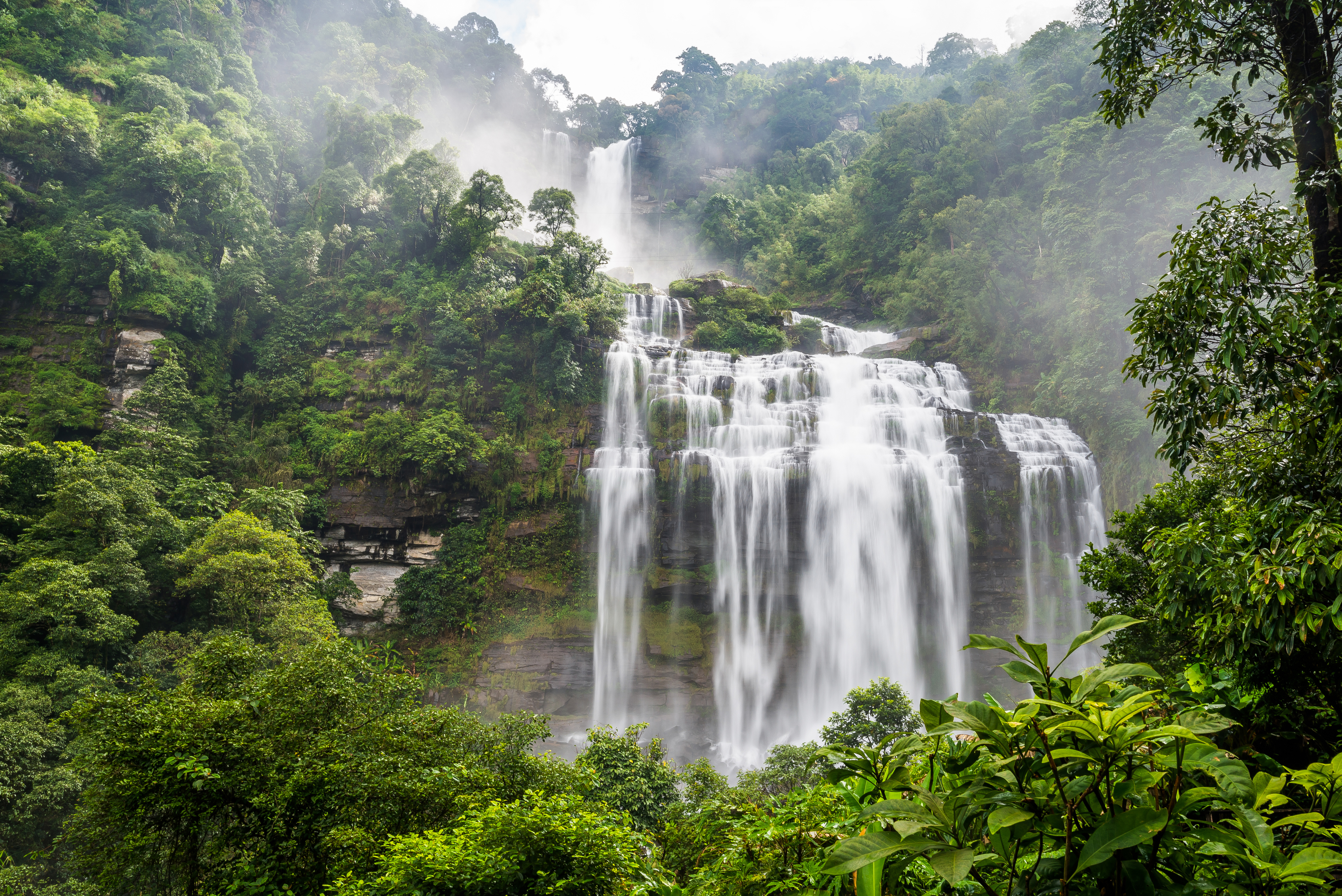 A tall waterfall drops through thick green jungle into a misty pool in Laos's Bolaven Plateau.