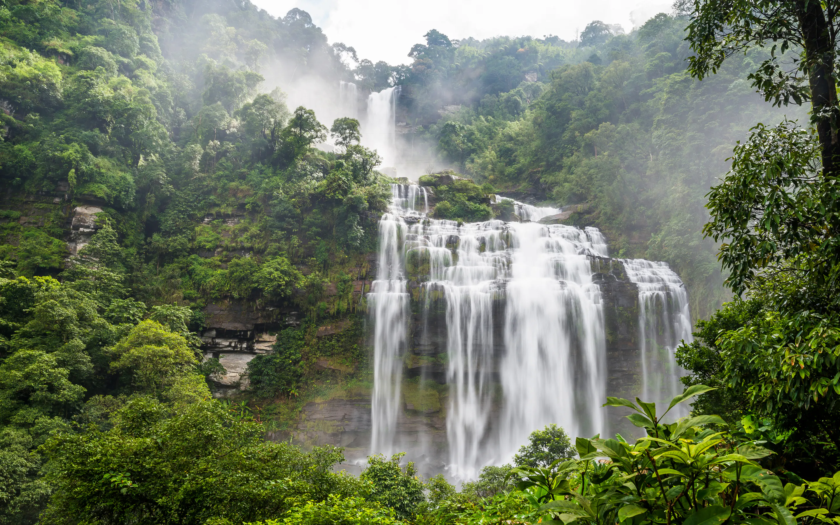 A tall waterfall drops through thick green jungle into a misty pool in Laos's Bolaven Plateau.