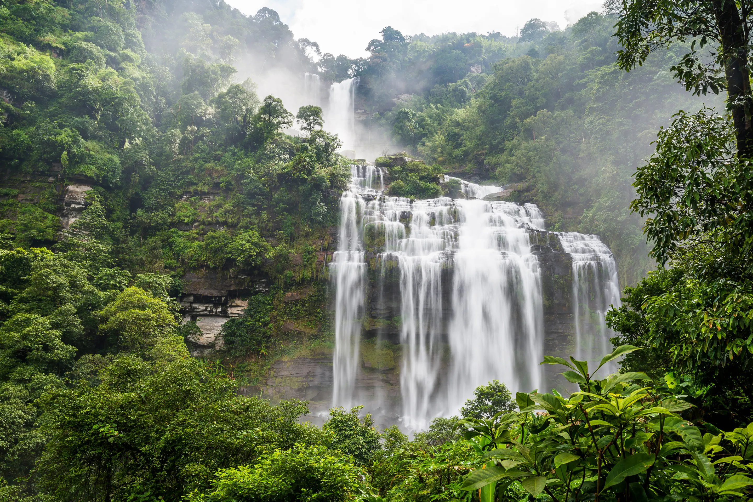 A tall waterfall drops through thick green jungle into a misty pool in Laos's Bolaven Plateau.