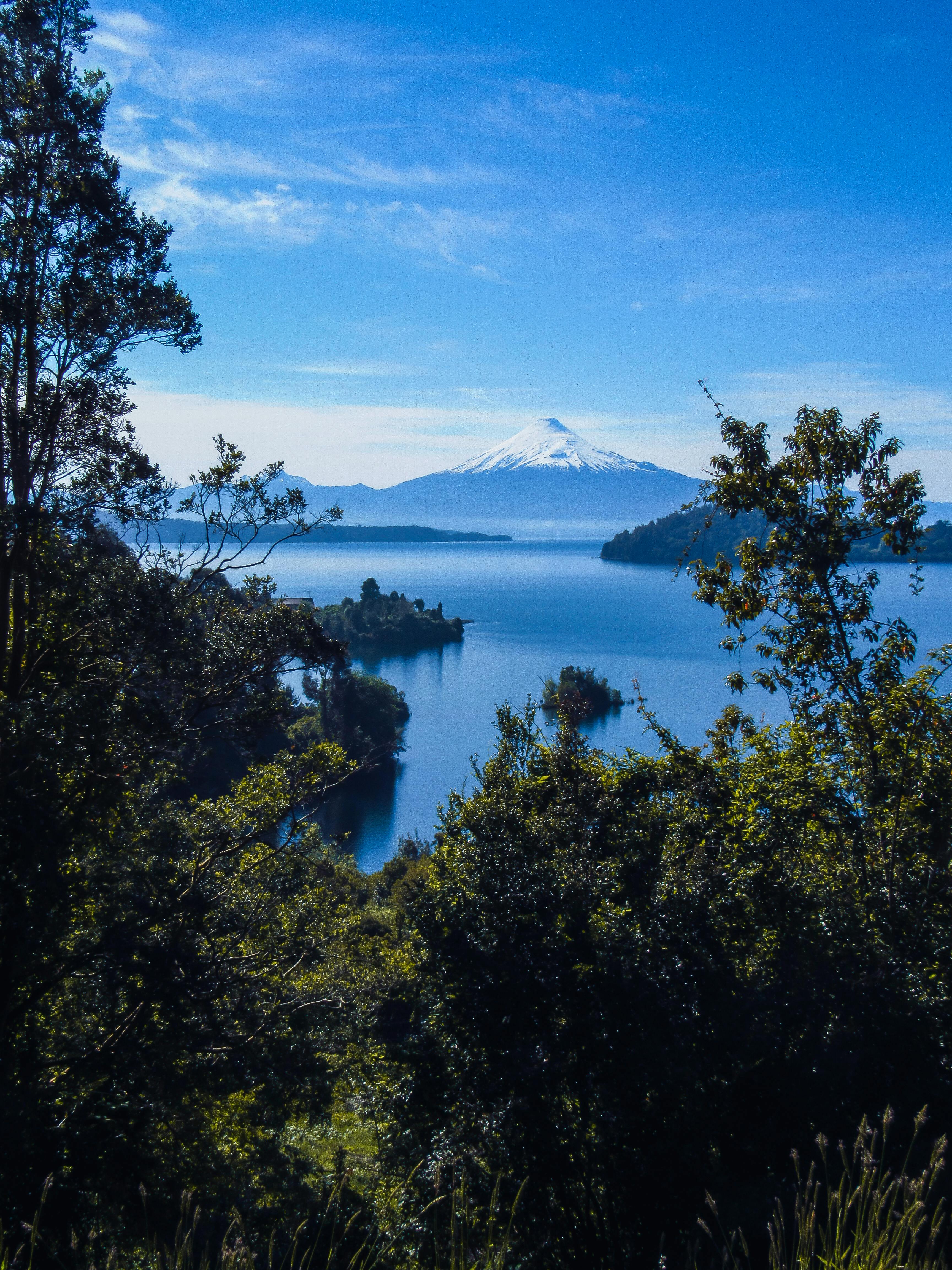 A placid lake reflects green forested hillsides and a distant snow-capped peak in Chile.