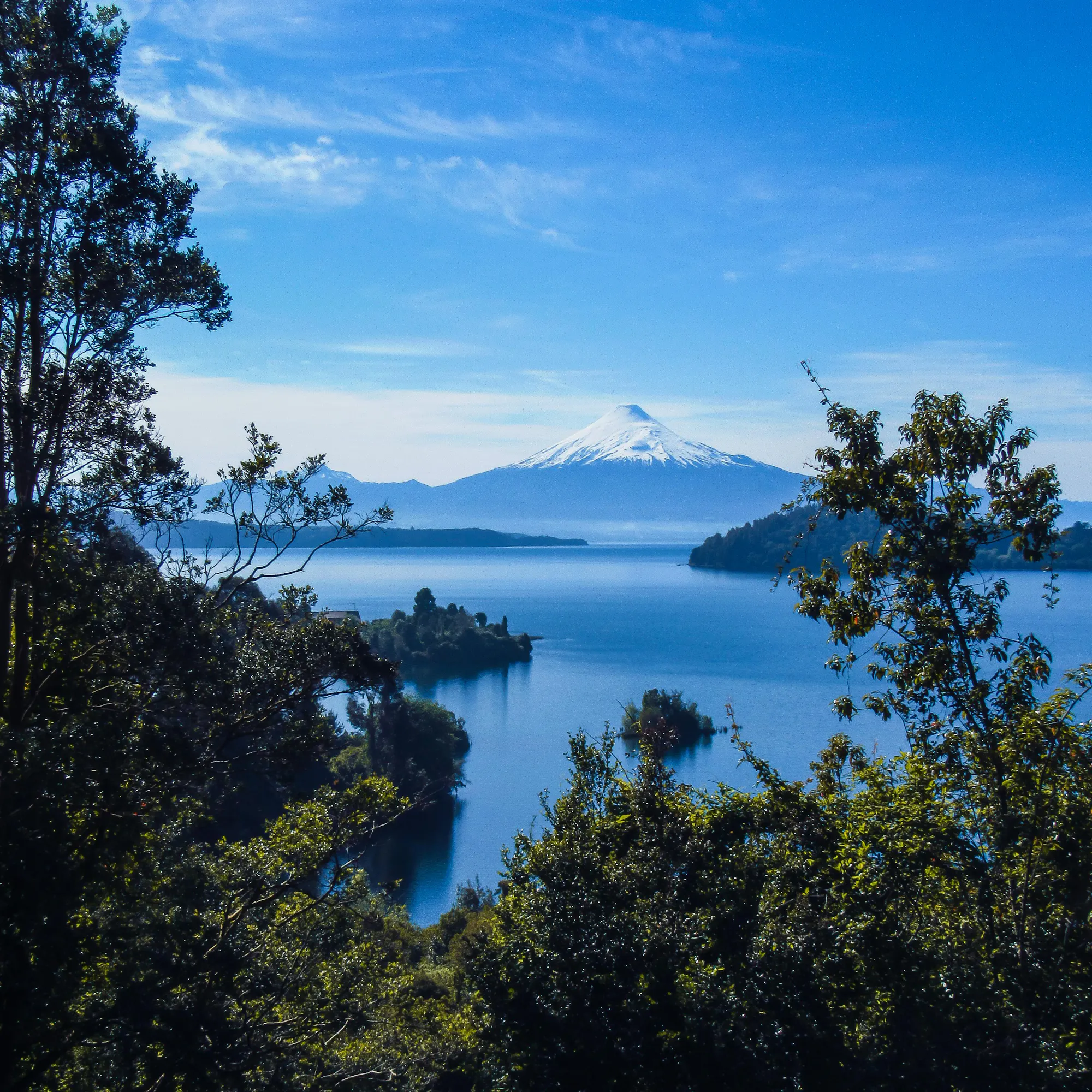 A placid lake reflects green forested hillsides and a distant snow-capped peak in Chile.