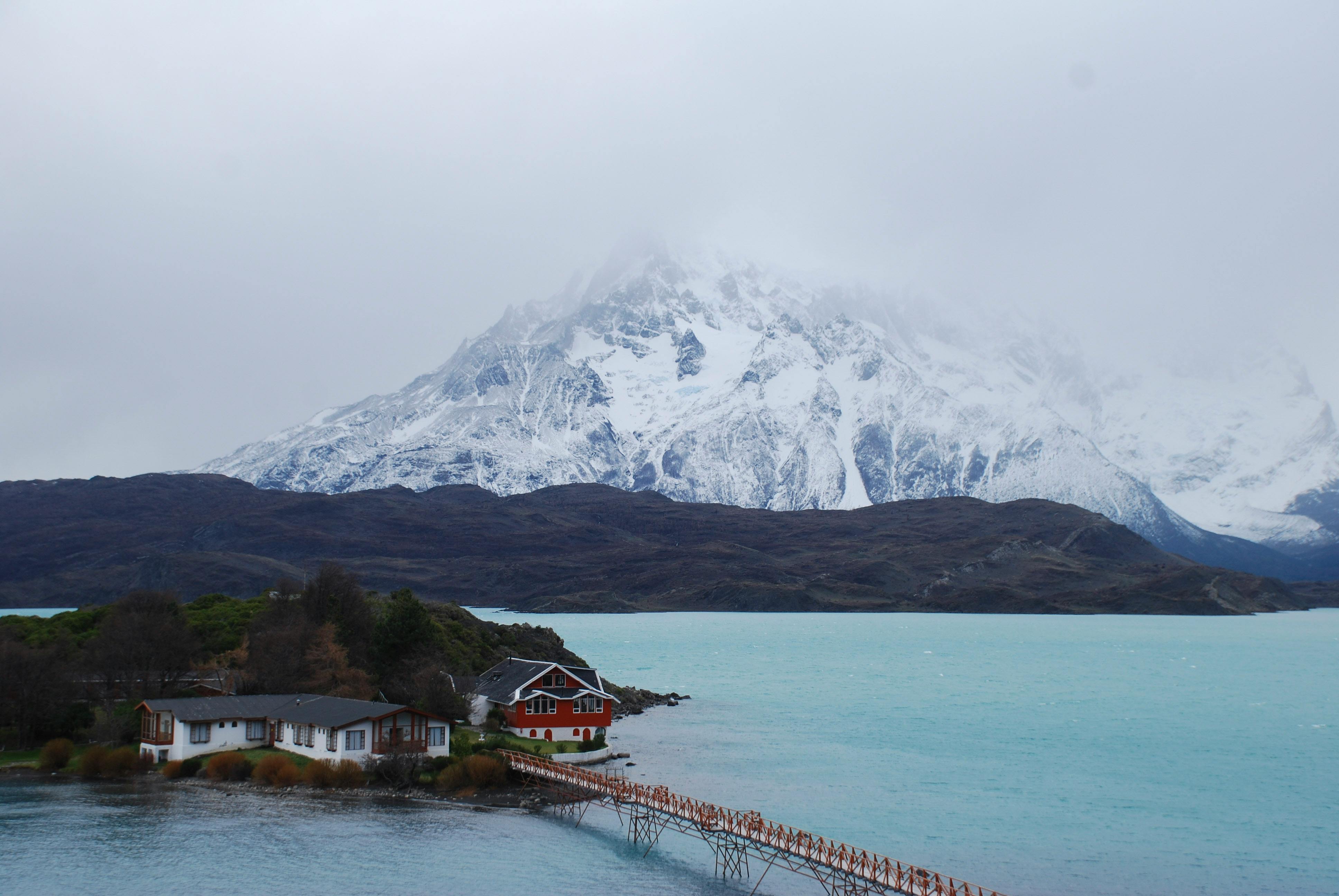 A snow-capped volcano rises behind a calm blue fjord lake with a small wooden dock in the foreground.