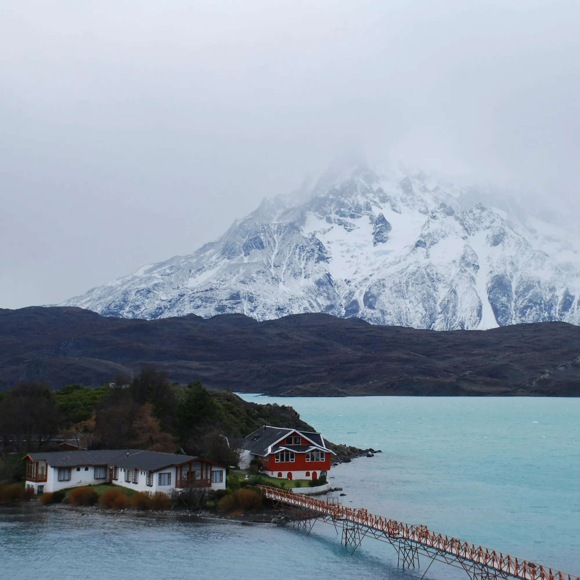 A snow-capped volcano rises behind a calm blue fjord lake with a small wooden dock in the foreground.