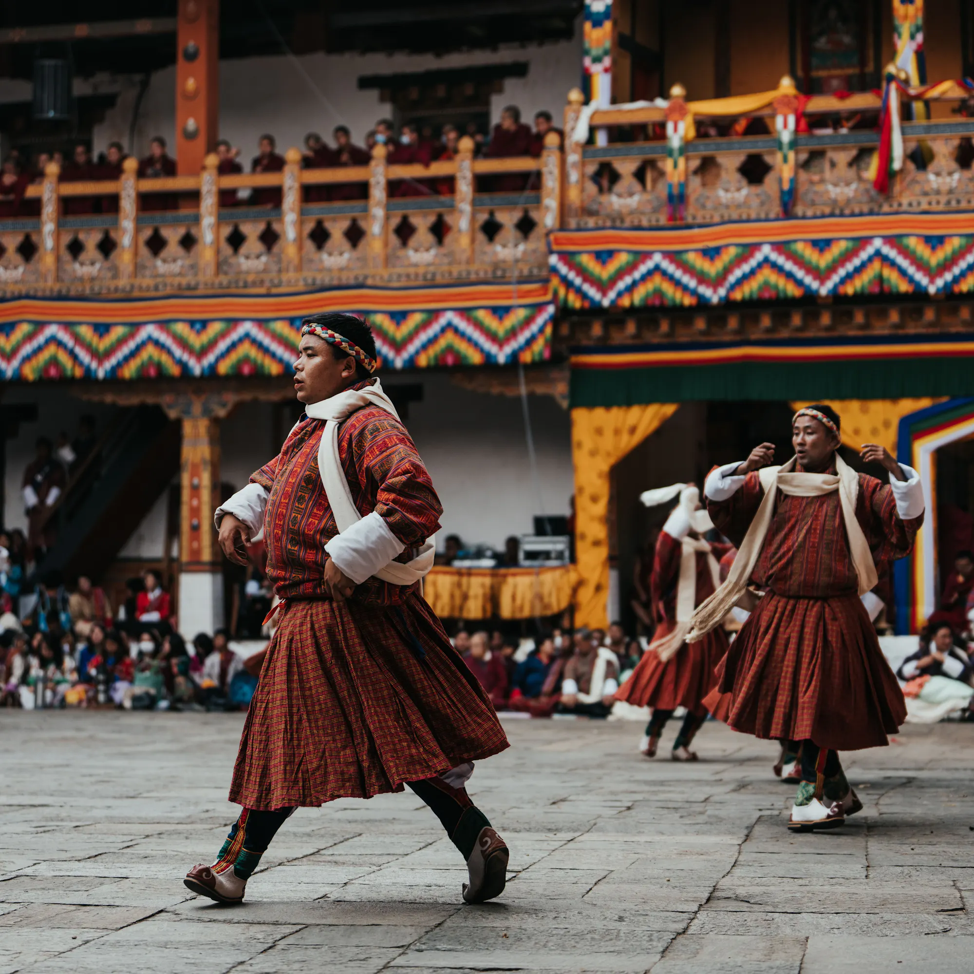 Monks in red and saffron robes perform a traditional dance in an ornate Bhutanese courtyard.