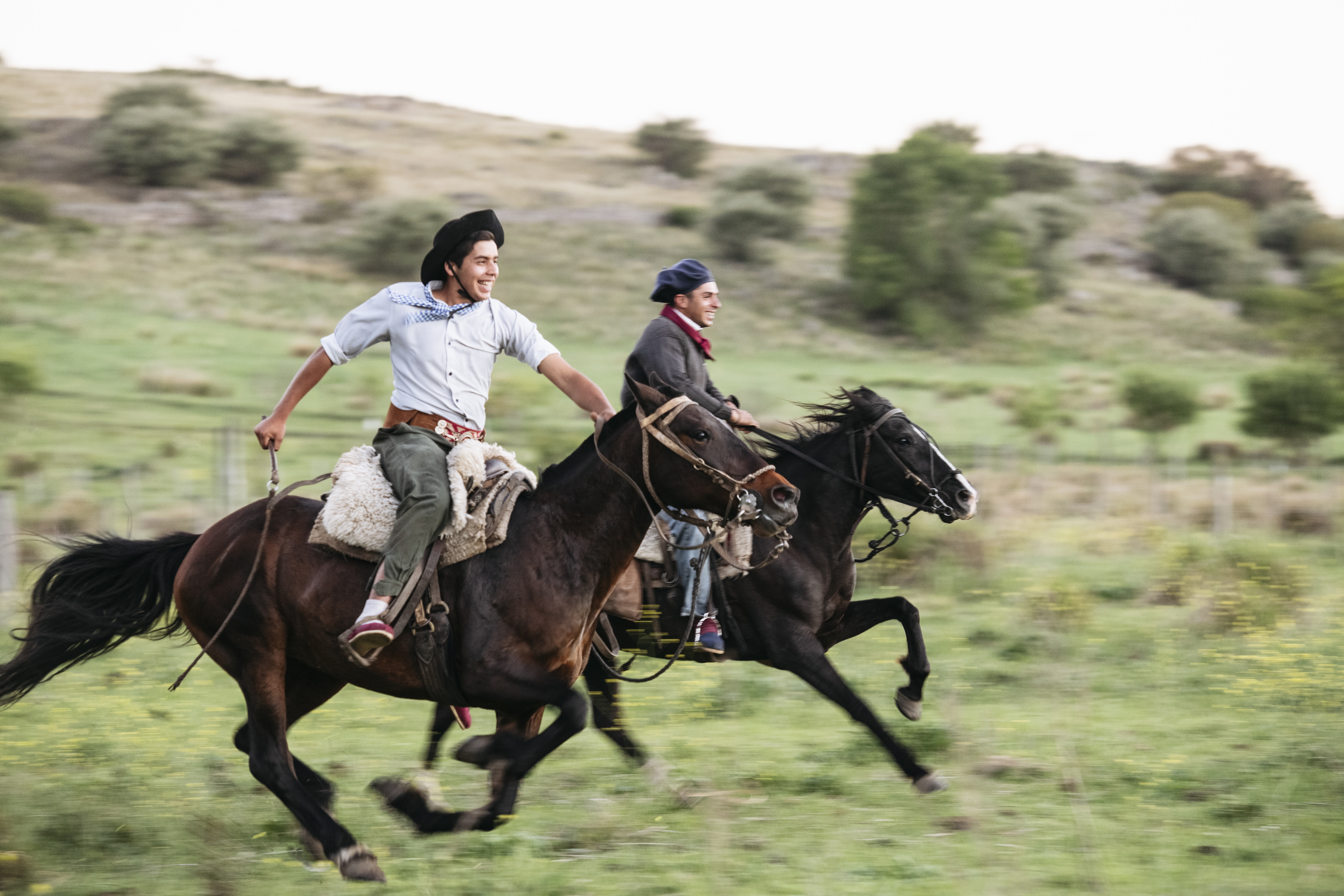 Two gauchos gallop on horseback across open green countryside in Córdoba province, Argentina.