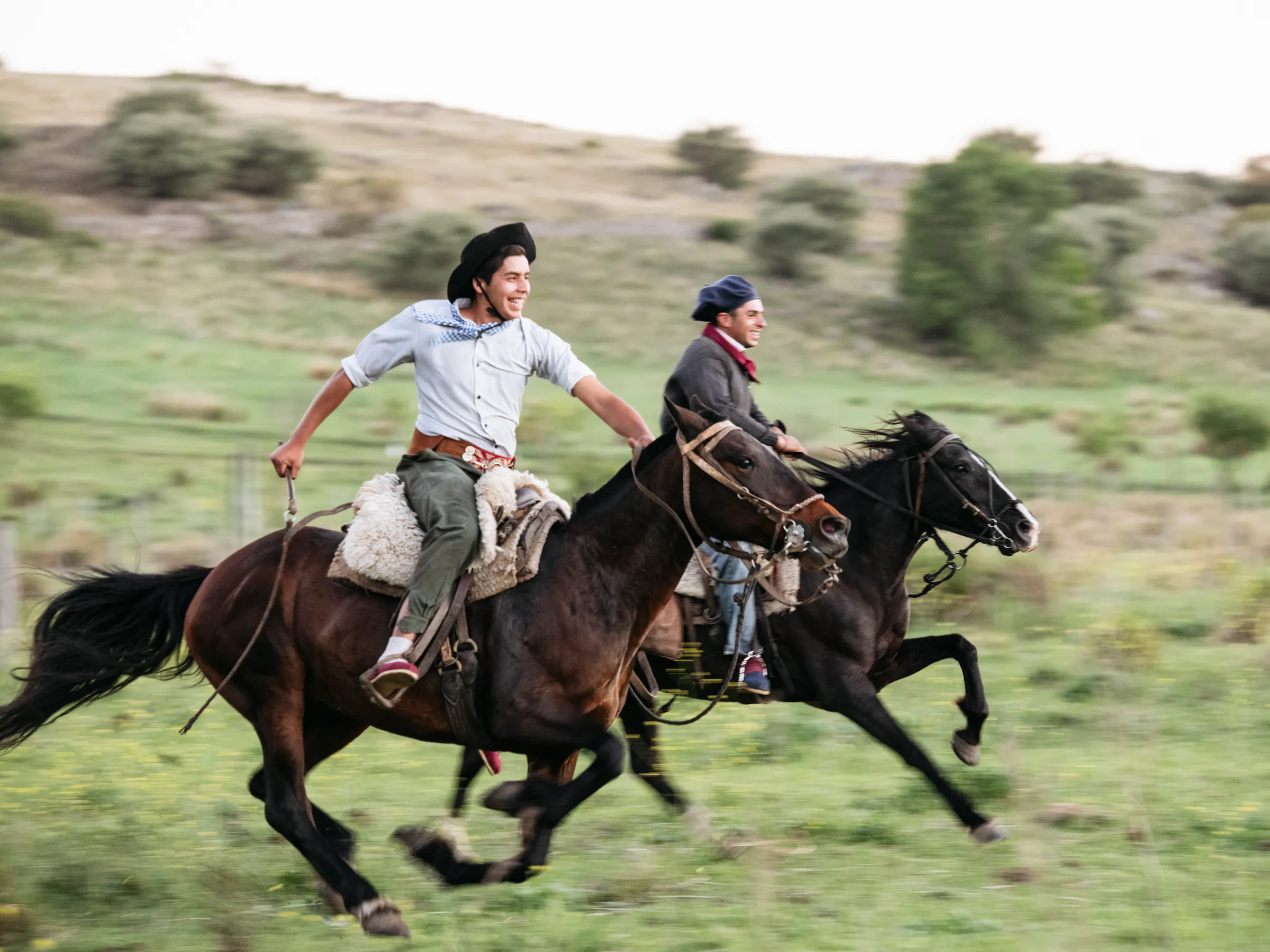 Two gauchos gallop on horseback across open green countryside in Córdoba province, Argentina.